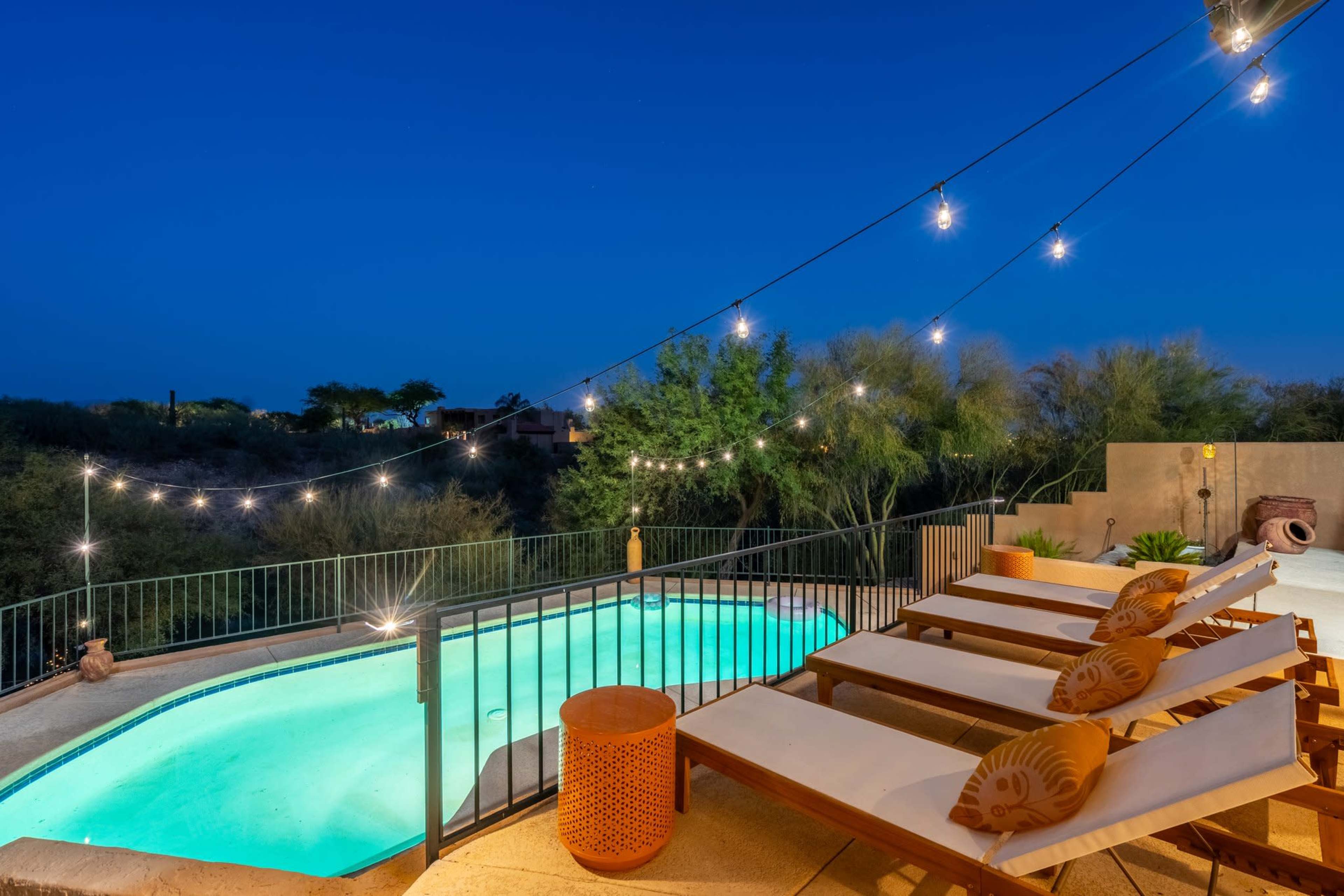 A patio area with lounge chairs and a swimming pool, illuminated by string lights at dusk.