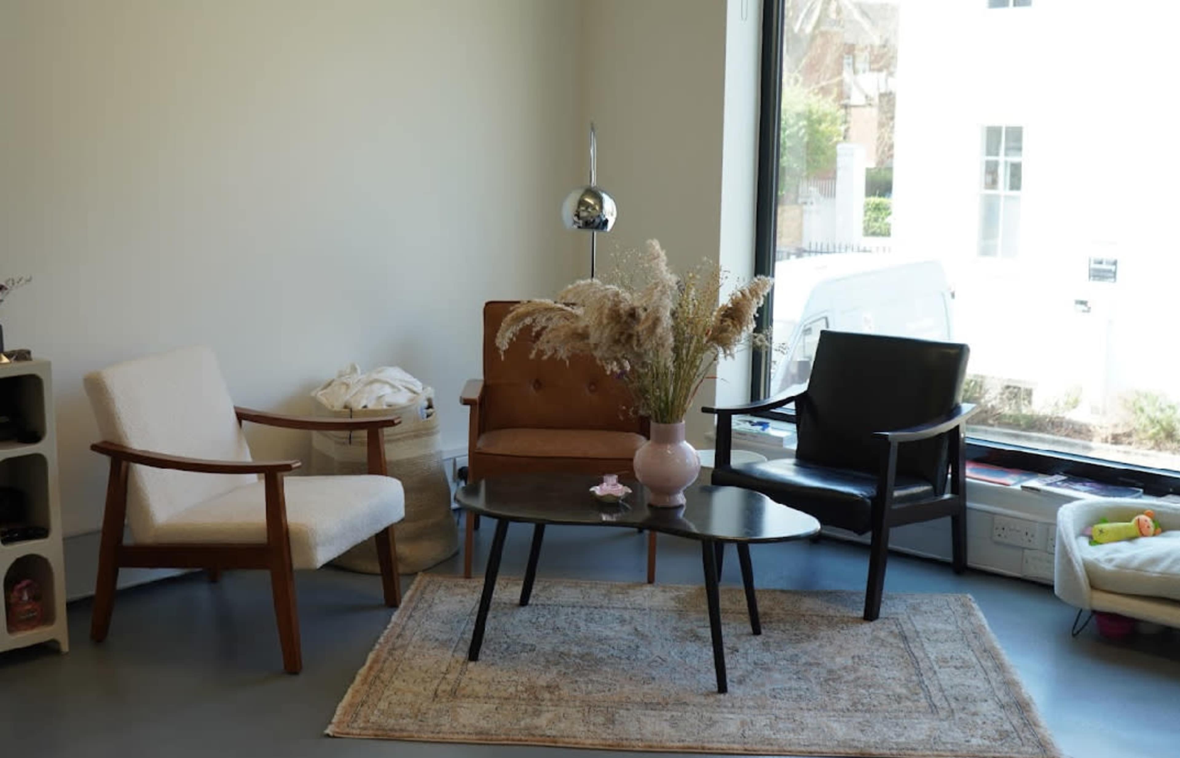 The image shows a modern seating area with two armchairs, a round coffee table with a vase of dried flowers, and a patterned rug in front of a large window.