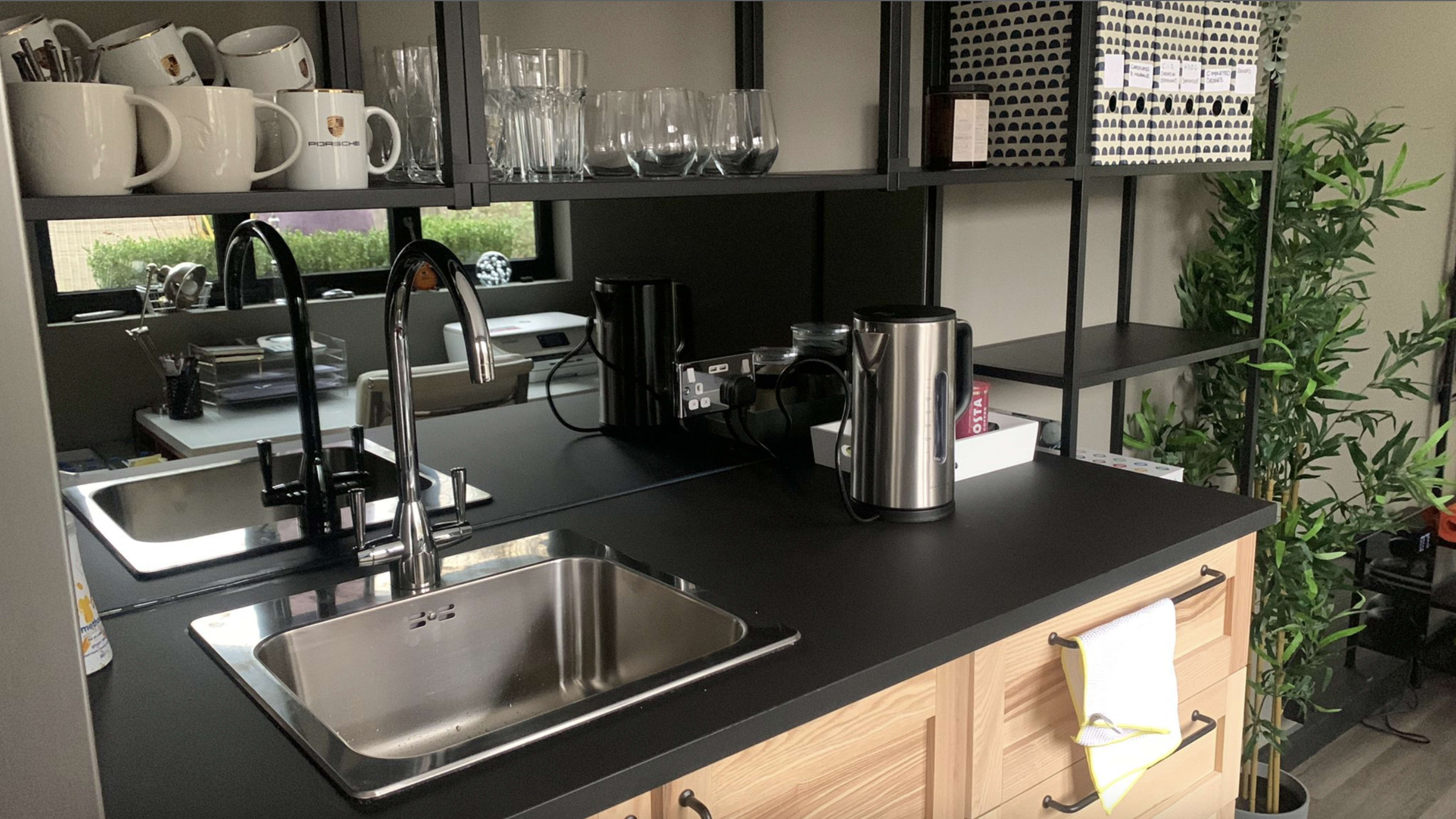 A modern kitchen with a stainless steel sink, a coffee machine, a kettle, and a variety of glassware arranged on open shelves.