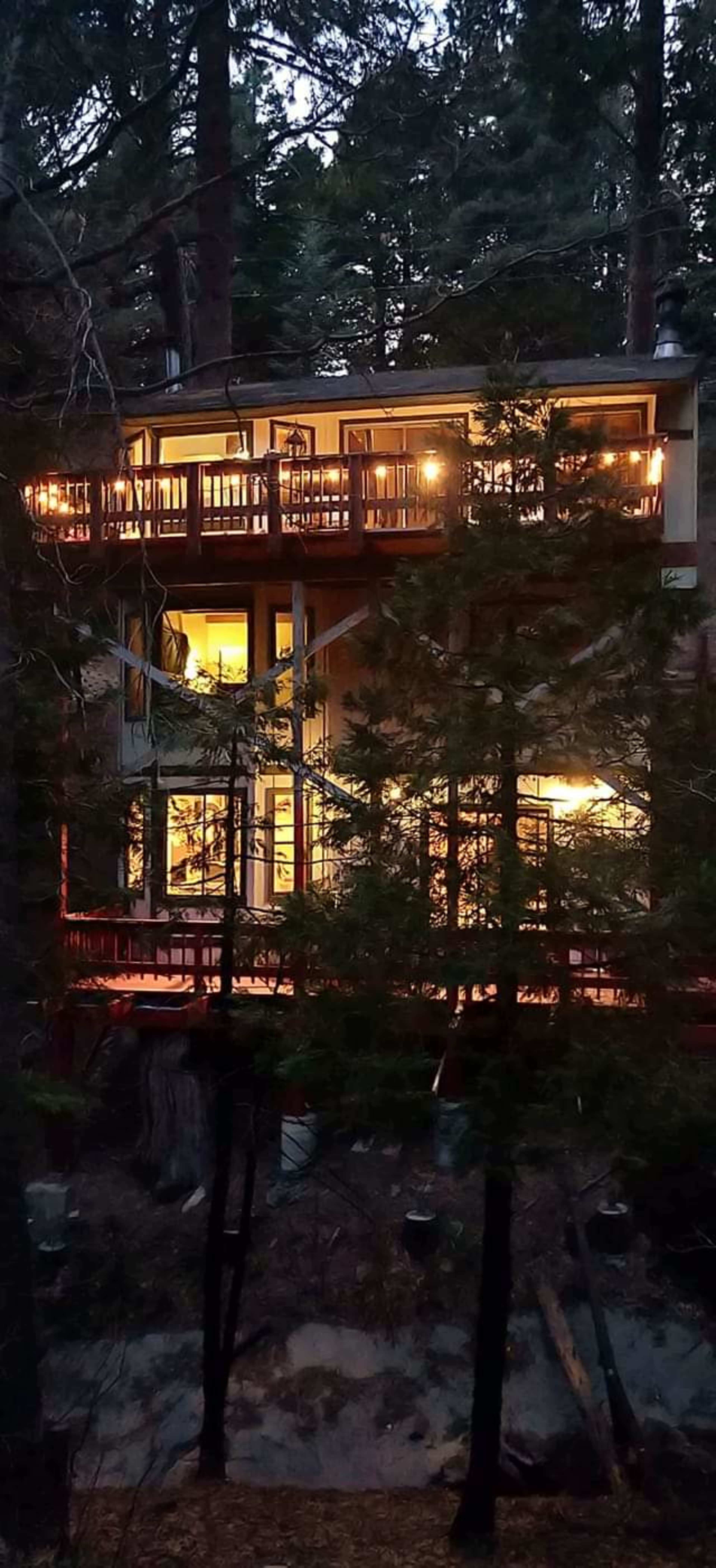 A two-story cabin with large windows and multiple balconies is nestled among tall trees in a forested area at dusk.