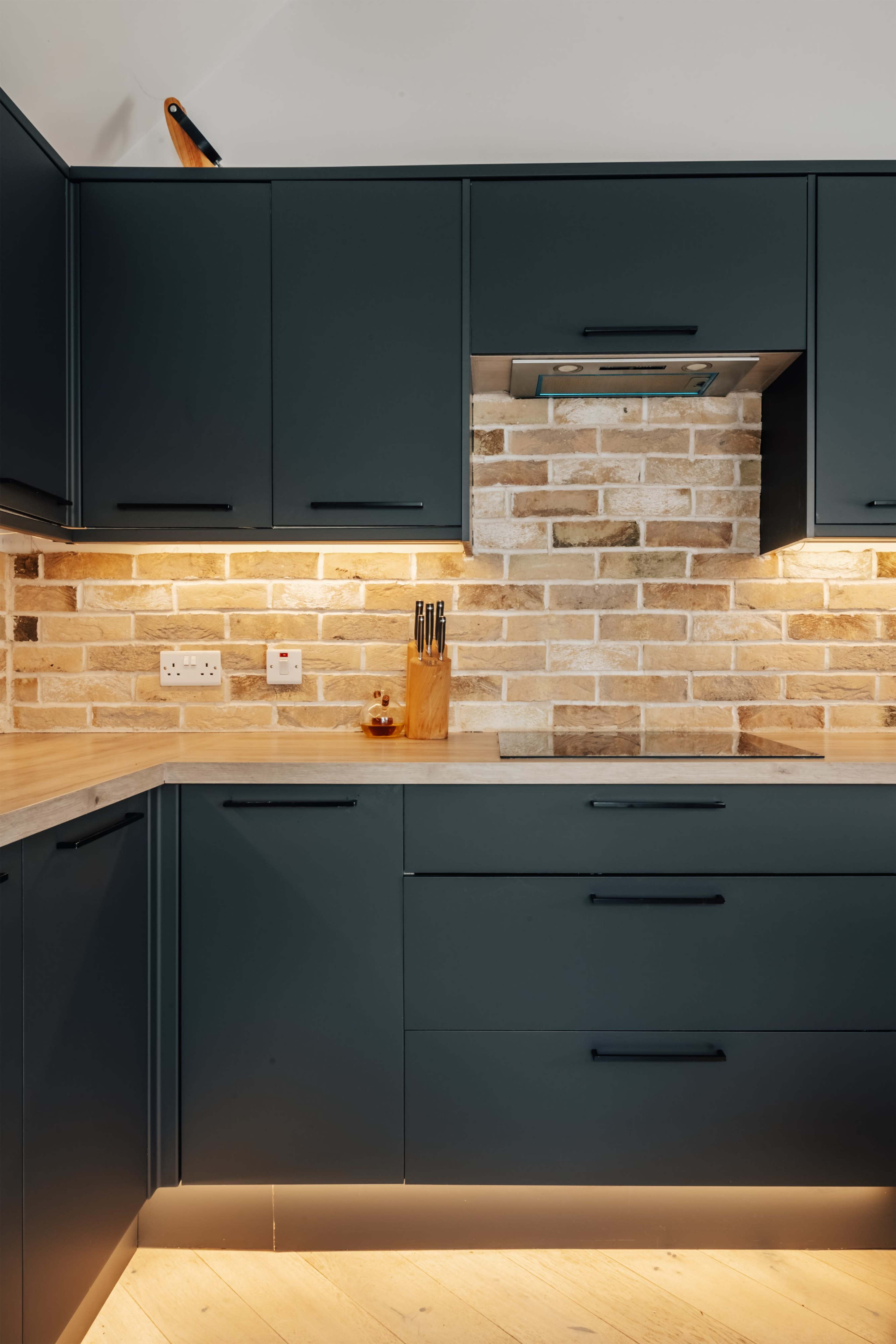 The image shows a modern kitchen with dark cabinetry, a light brick backsplash, and under-cabinet lighting illuminating the countertop.