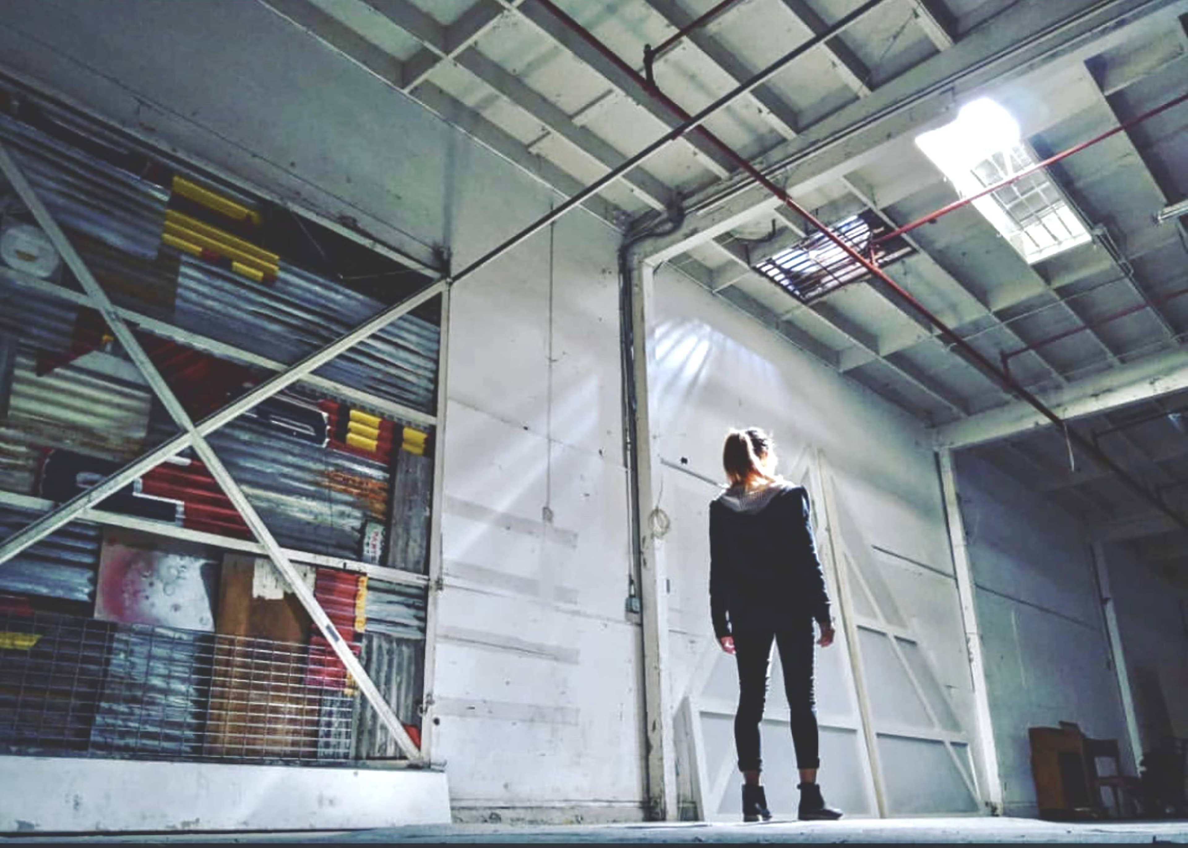 A person stands in an empty warehouse with a high ceiling and light streaming through a skylight.