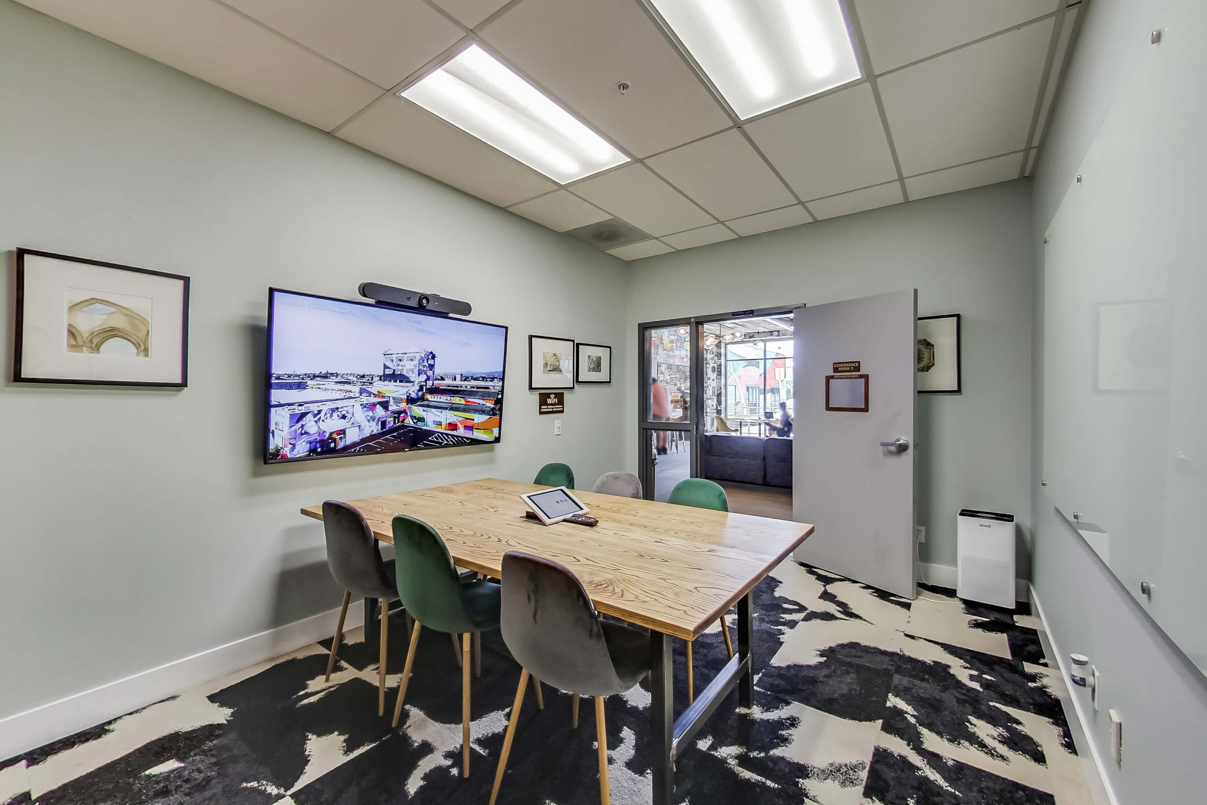The image shows a small conference room featuring a wooden table with chairs, a large wall-mounted screen, and an open door leading to an adjacent space.