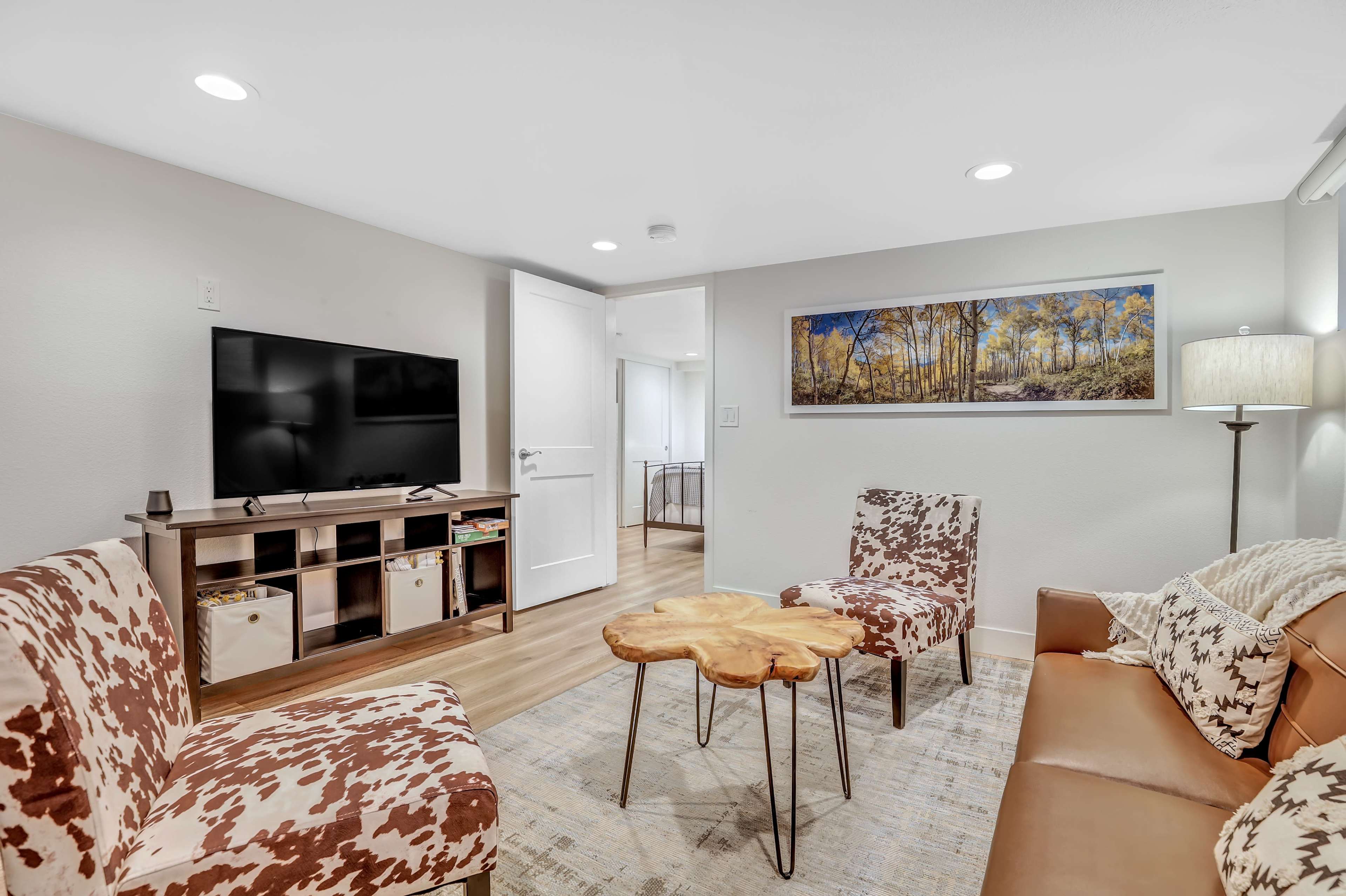 The room features a television mounted on the wall, a wooden coffee table, two upholstered chairs with a cow print pattern, and a couch, all arranged in a cozy layout.