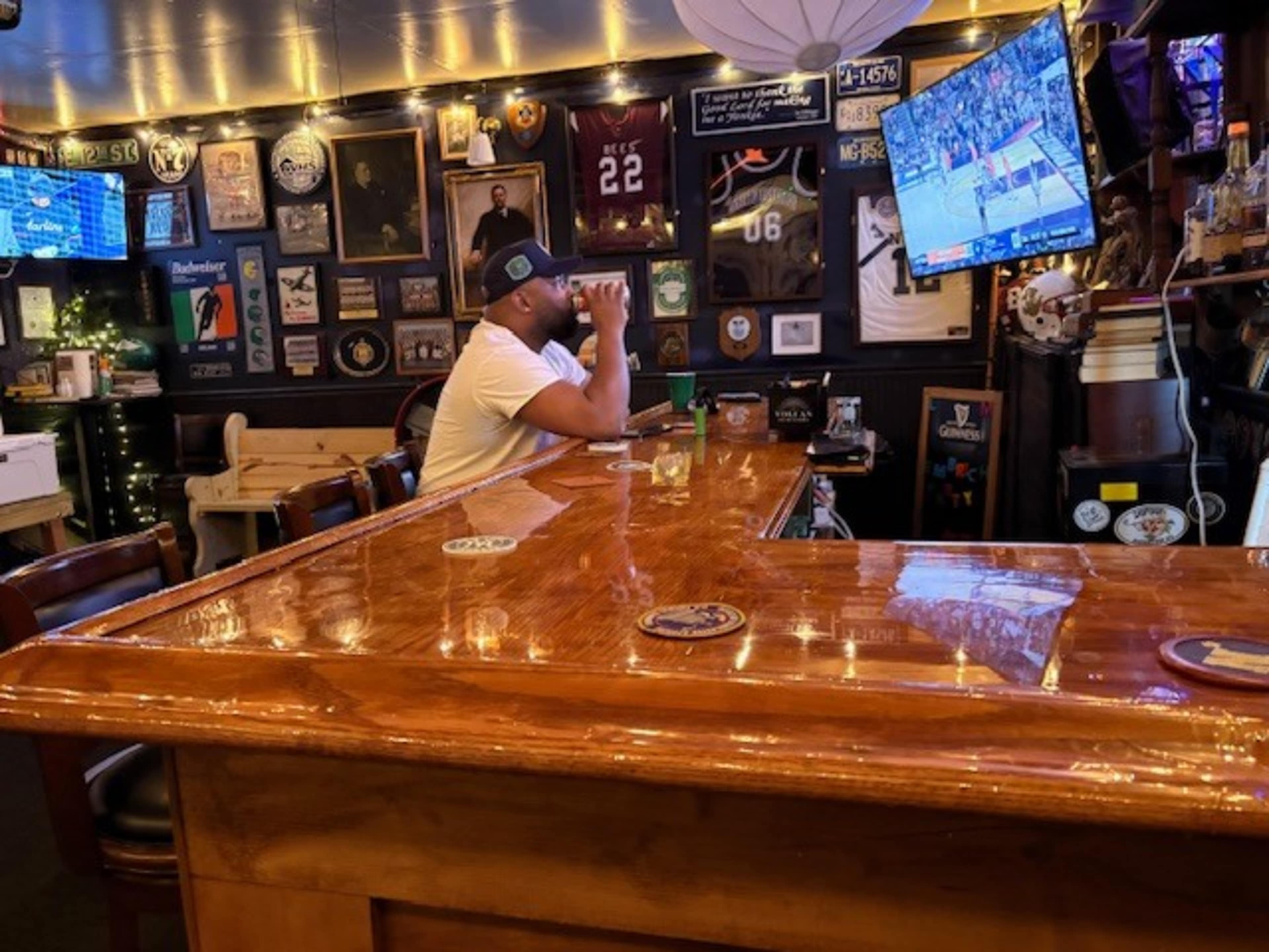 A man sits alone at a bar, watching a basketball game on a television, surrounded by framed memorabilia and various bar decorations.