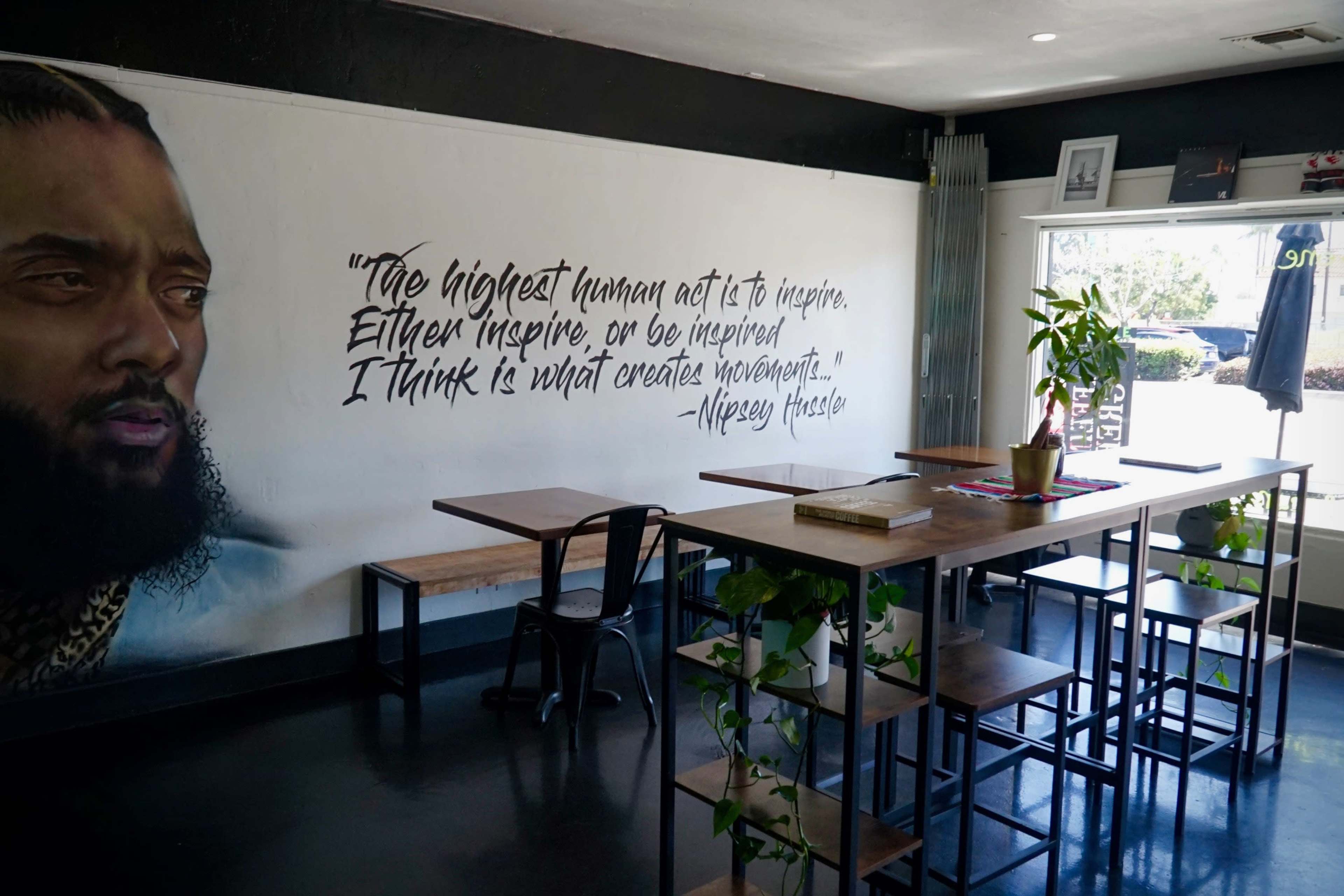 A minimalist café interior with a large mural of a man alongside a quote about inspiration, featuring wooden tables and metal chairs.