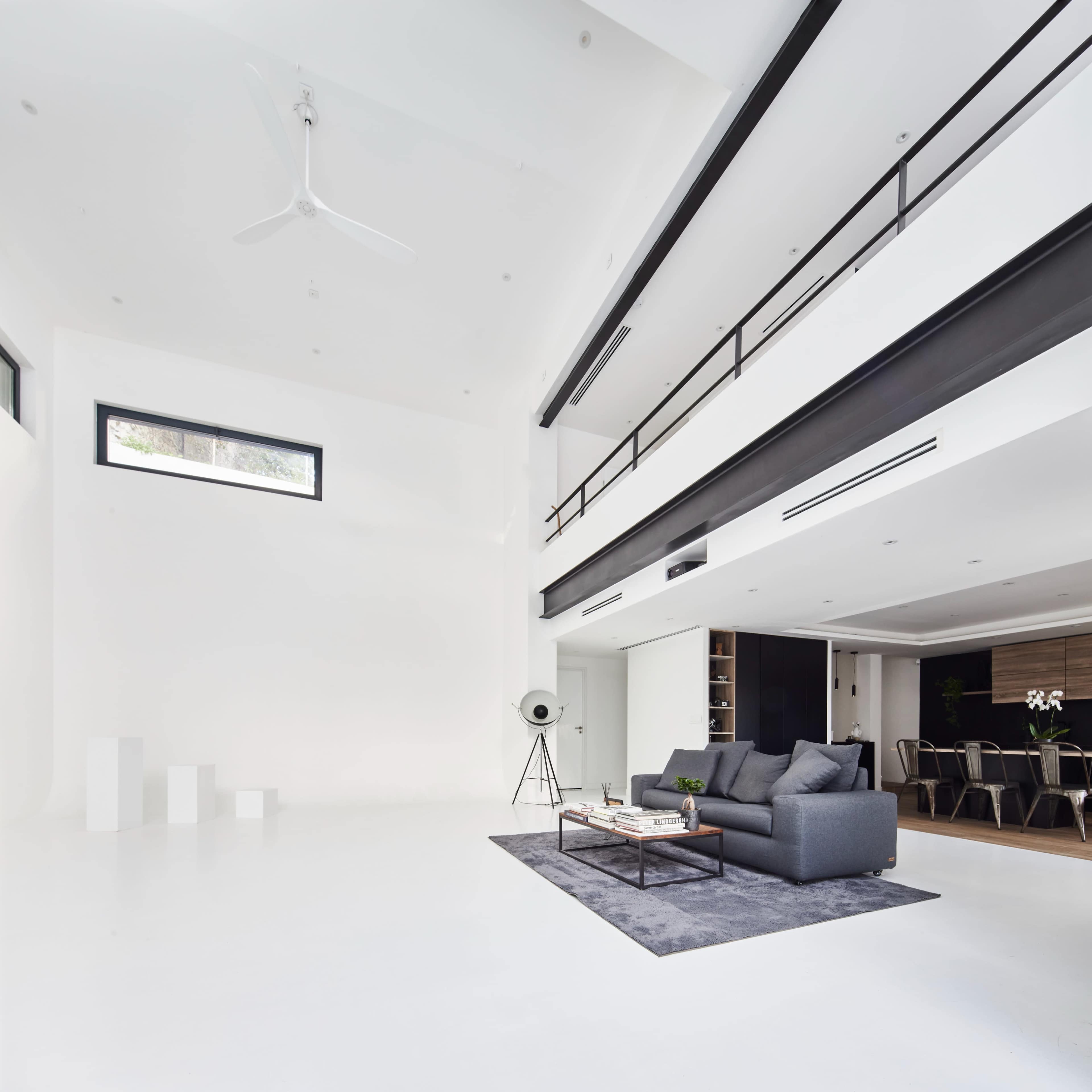 A modern, minimalist living space featuring a gray sofa, a coffee table, and a large rug on a white floor with high ceilings and an open layout.