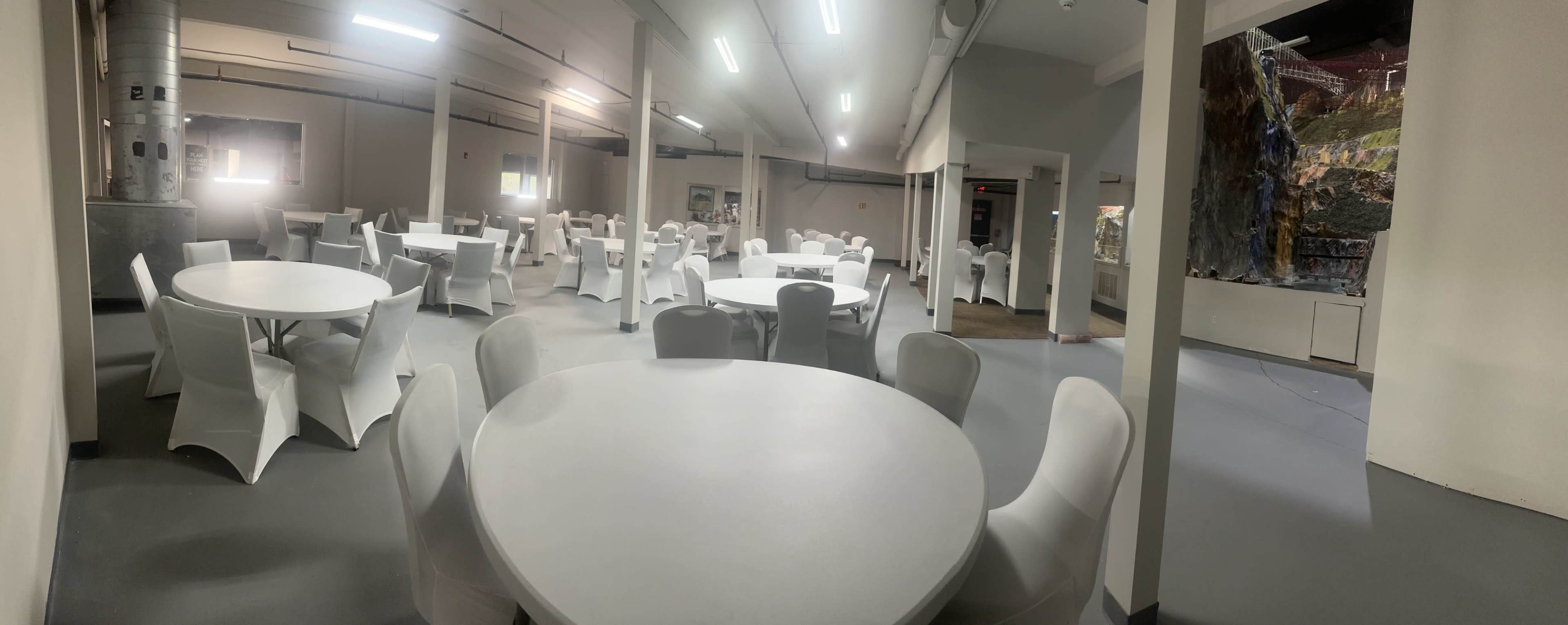 The image shows an empty room with several round tables surrounded by white chairs, illuminated by artificial lighting.