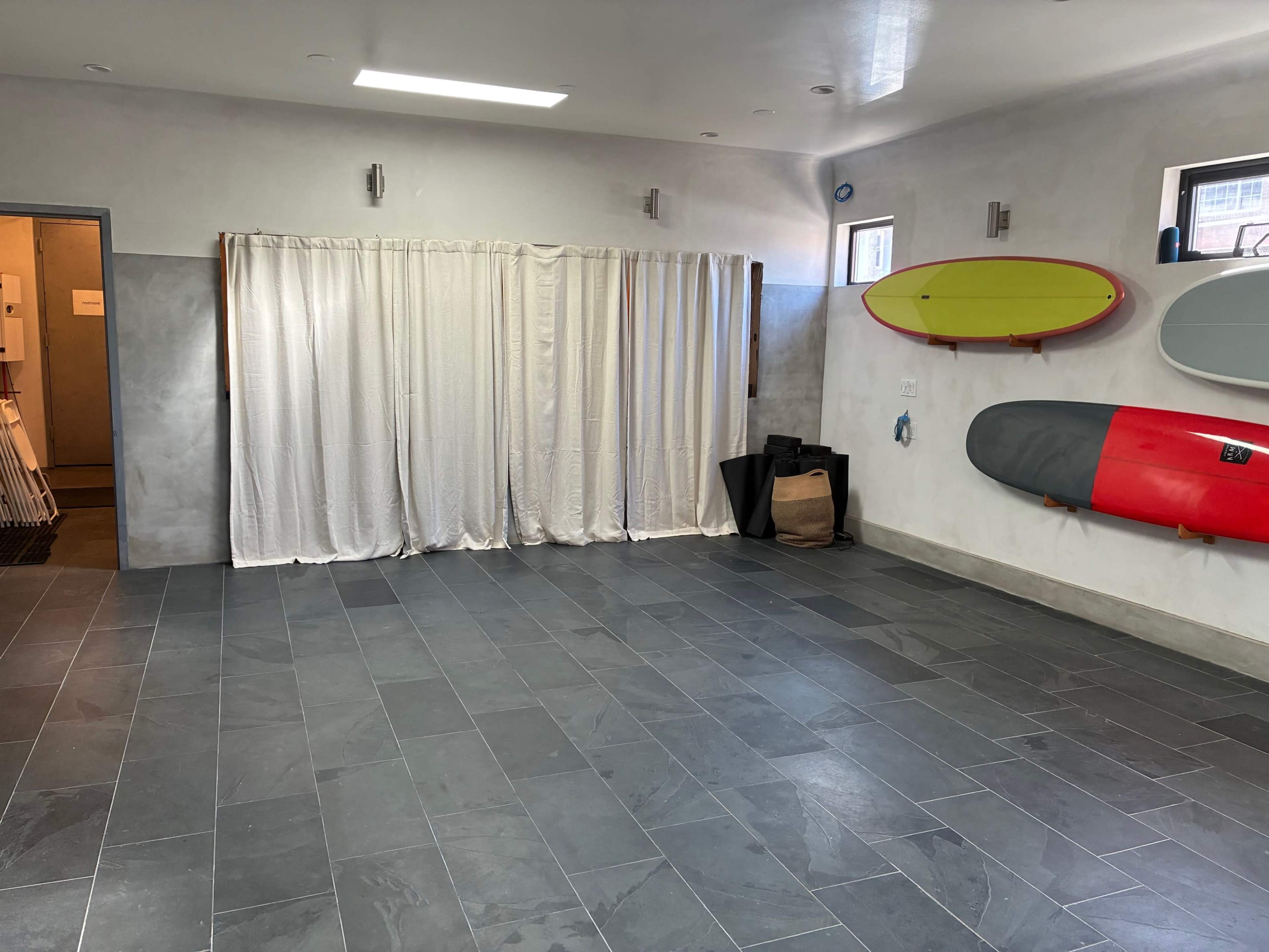 The image shows a spacious, minimalist room with gray tile flooring, draped white curtains on one side, and several surfboards mounted on the walls.