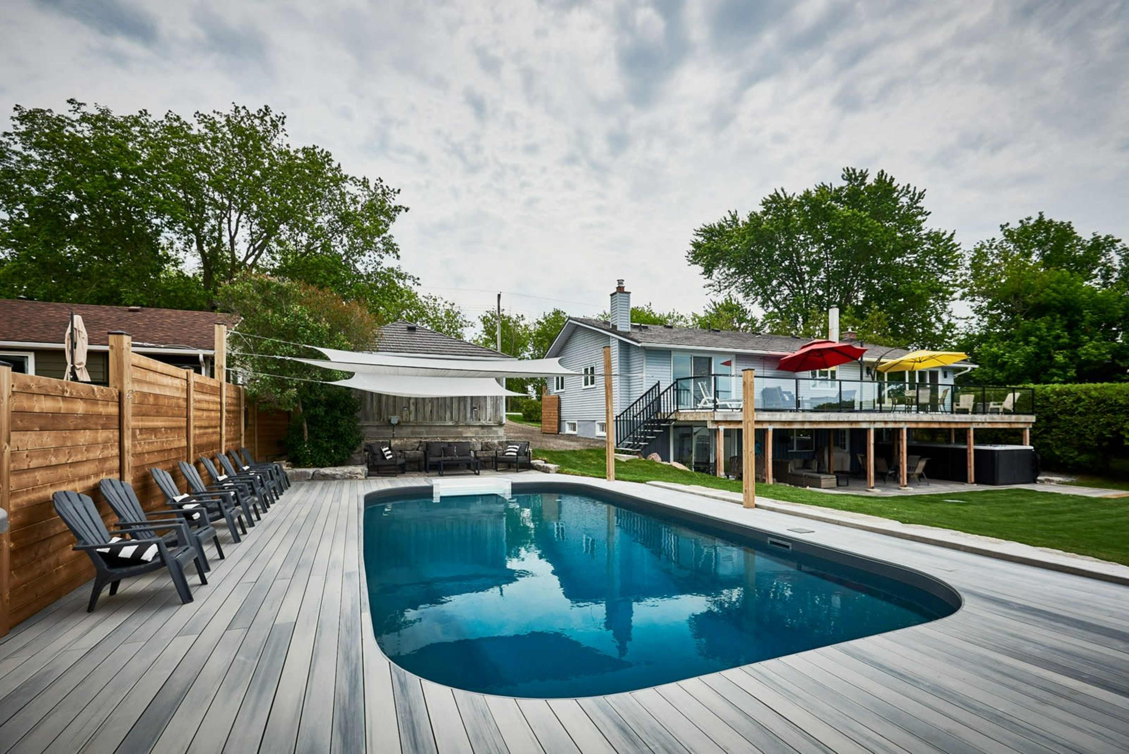 A backyard with a rectangular pool surrounded by wooden decking, lounge chairs, and a house with a deck and colorful umbrellas.