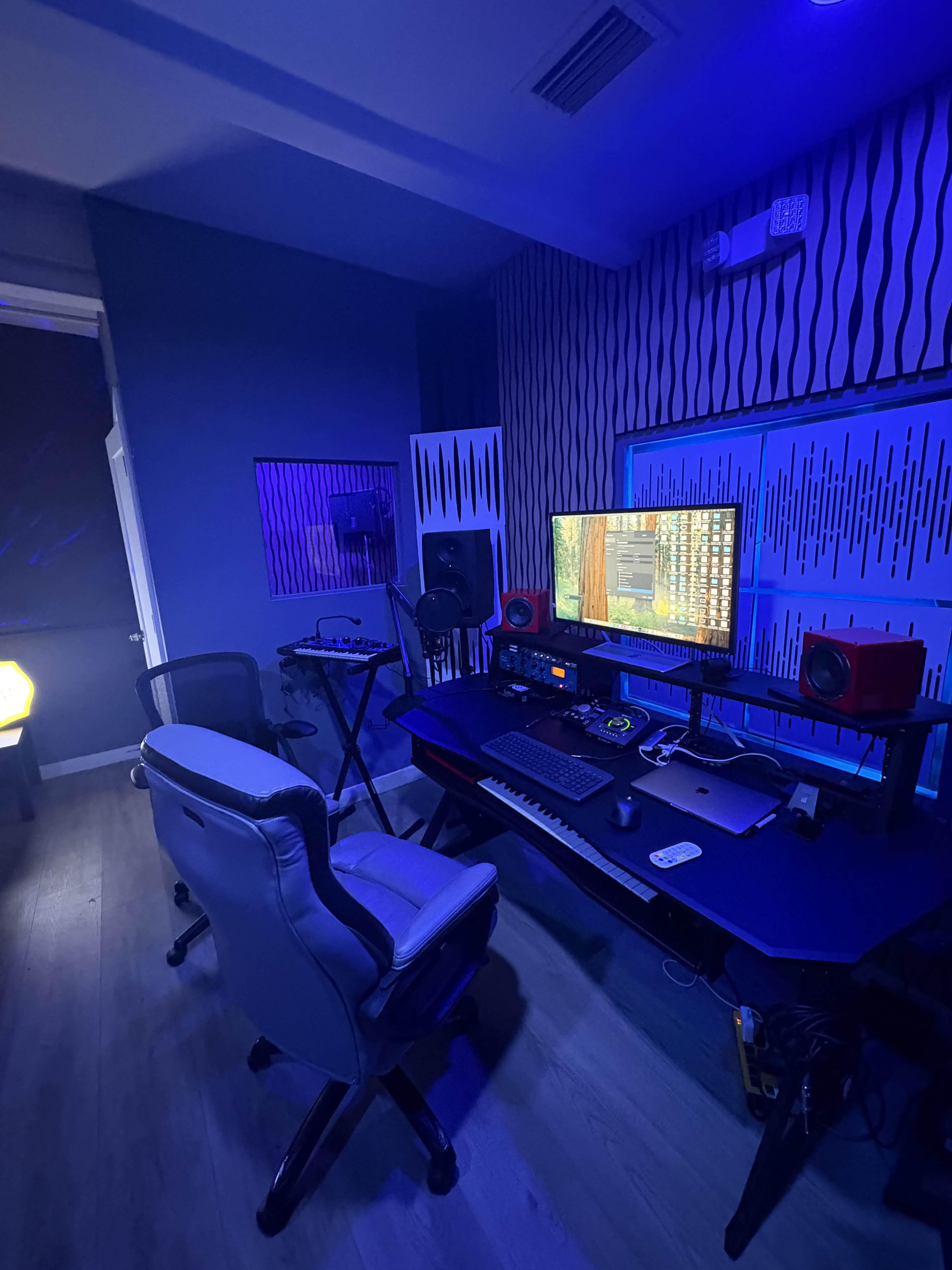 The image shows a music production studio with a desk featuring a computer, speakers, and various equipment, illuminated by blue lighting.