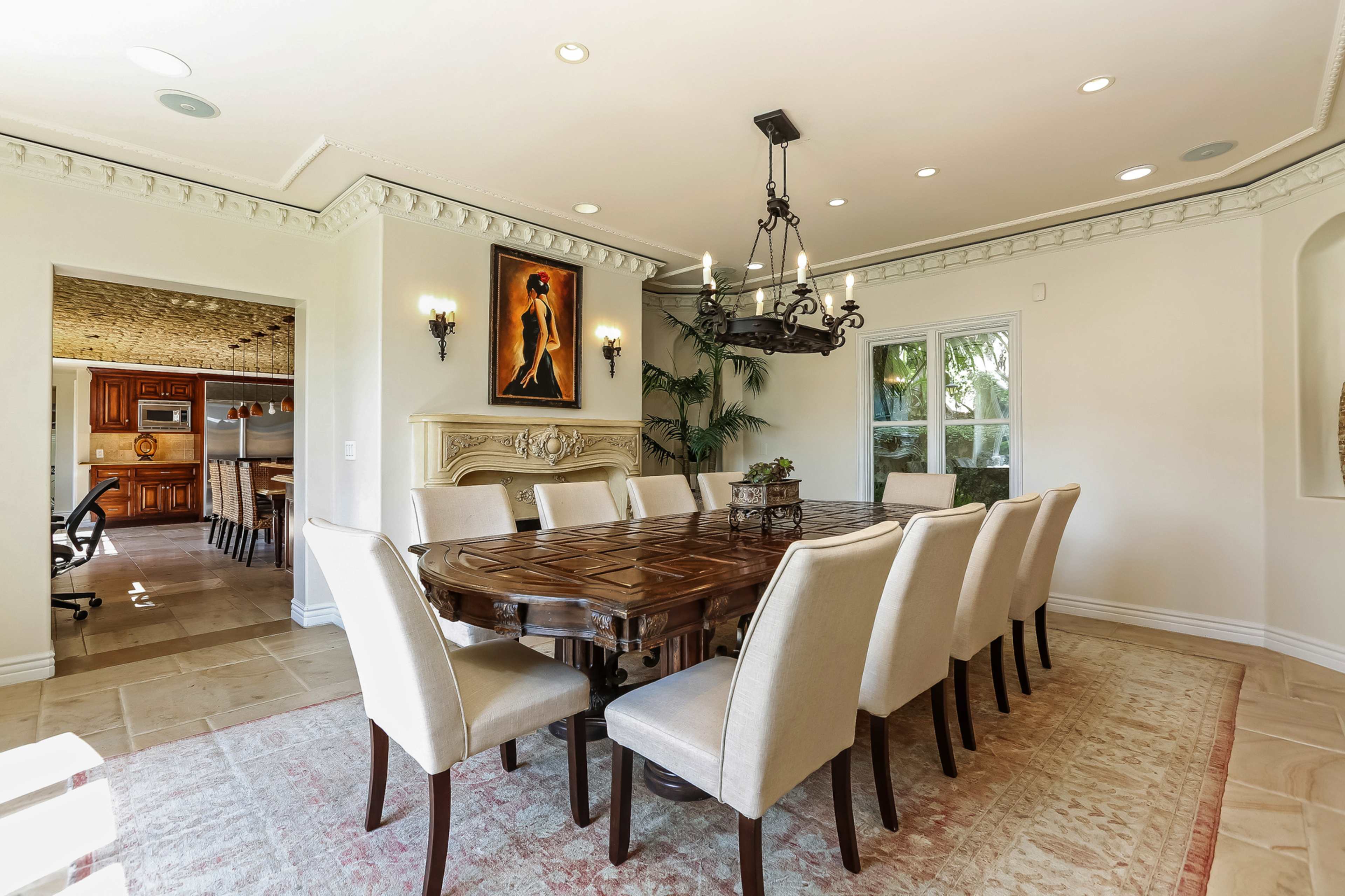A large dining room features a wooden table surrounded by twelve beige chairs, with a chandelier hanging overhead and a painting on the wall.