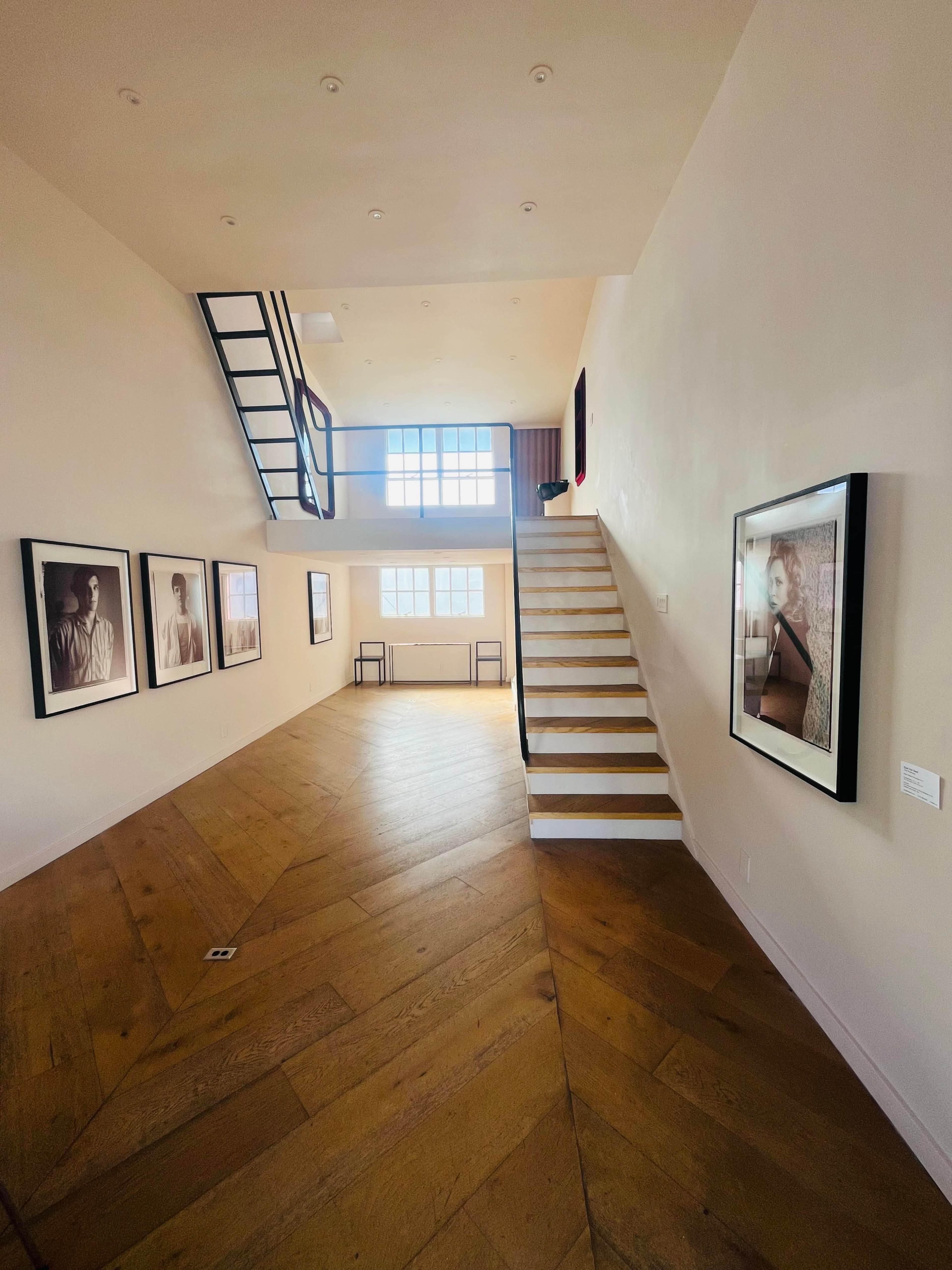 The image shows a spacious gallery with wooden flooring, featuring portraits on the walls and a staircase leading to the upper level.