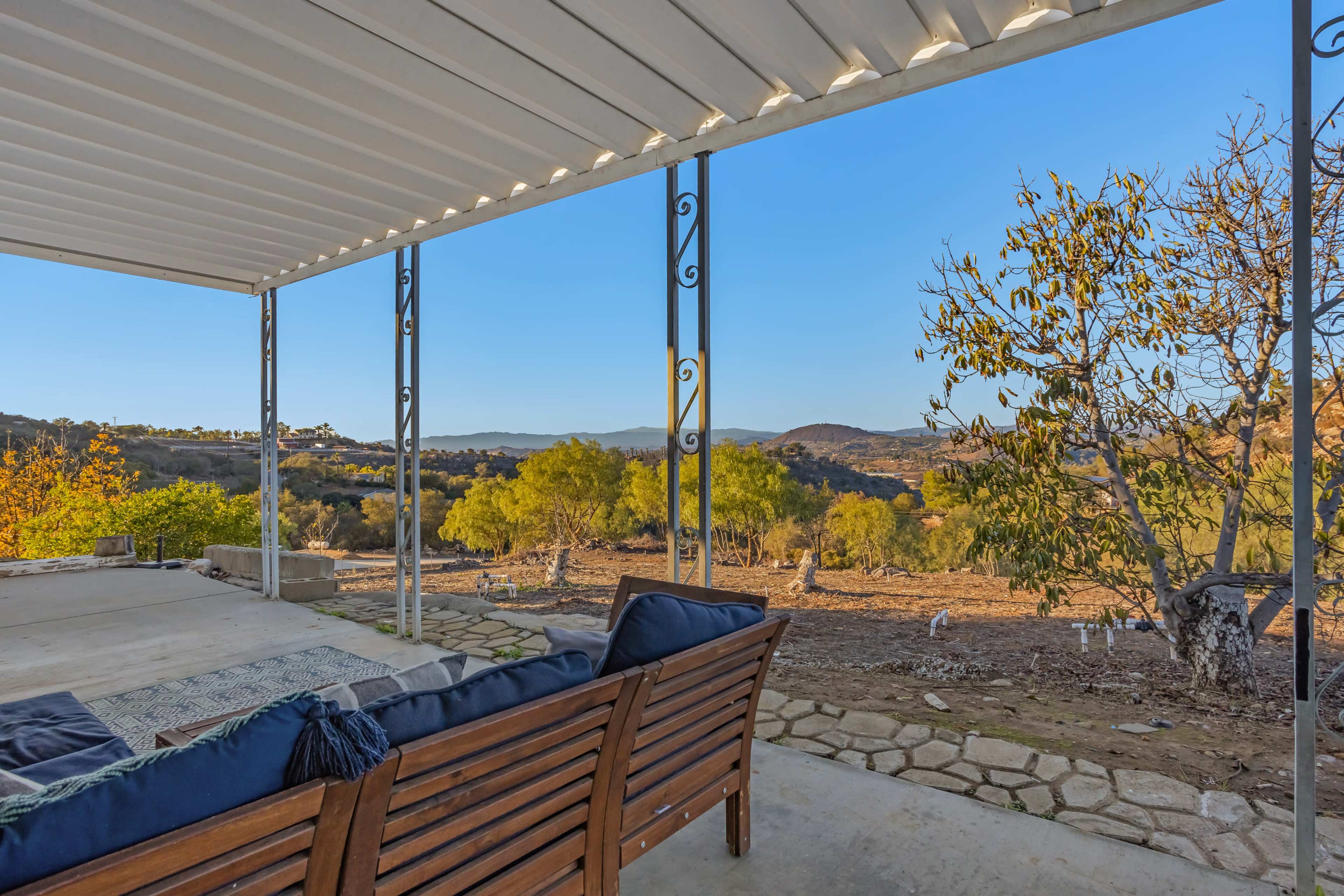 A covered patio with wooden seating overlooks a hilly landscape filled with trees under a clear blue sky.