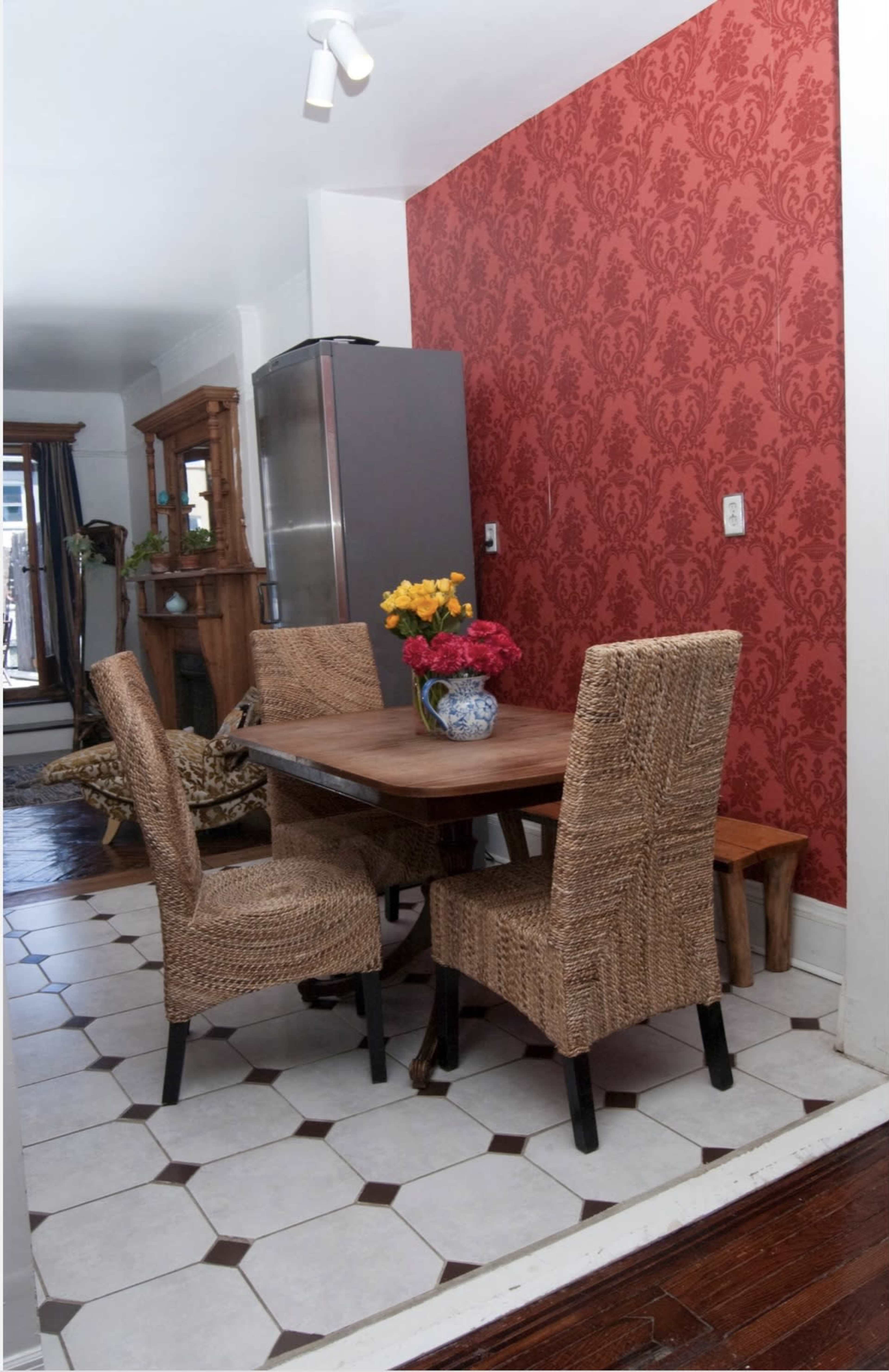 A dining area features a wooden table surrounded by woven chairs, with a floral centerpiece and a red patterned wallpaper in the background.
