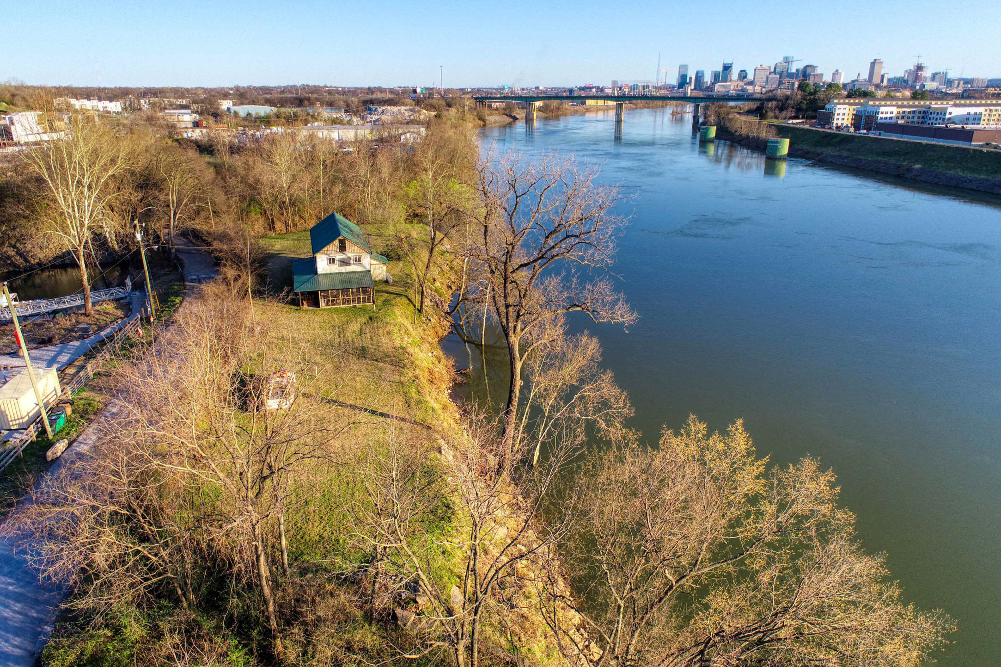 A river flows past a green-roofed house on a wooded bank, with a city skyline visible in the background.
