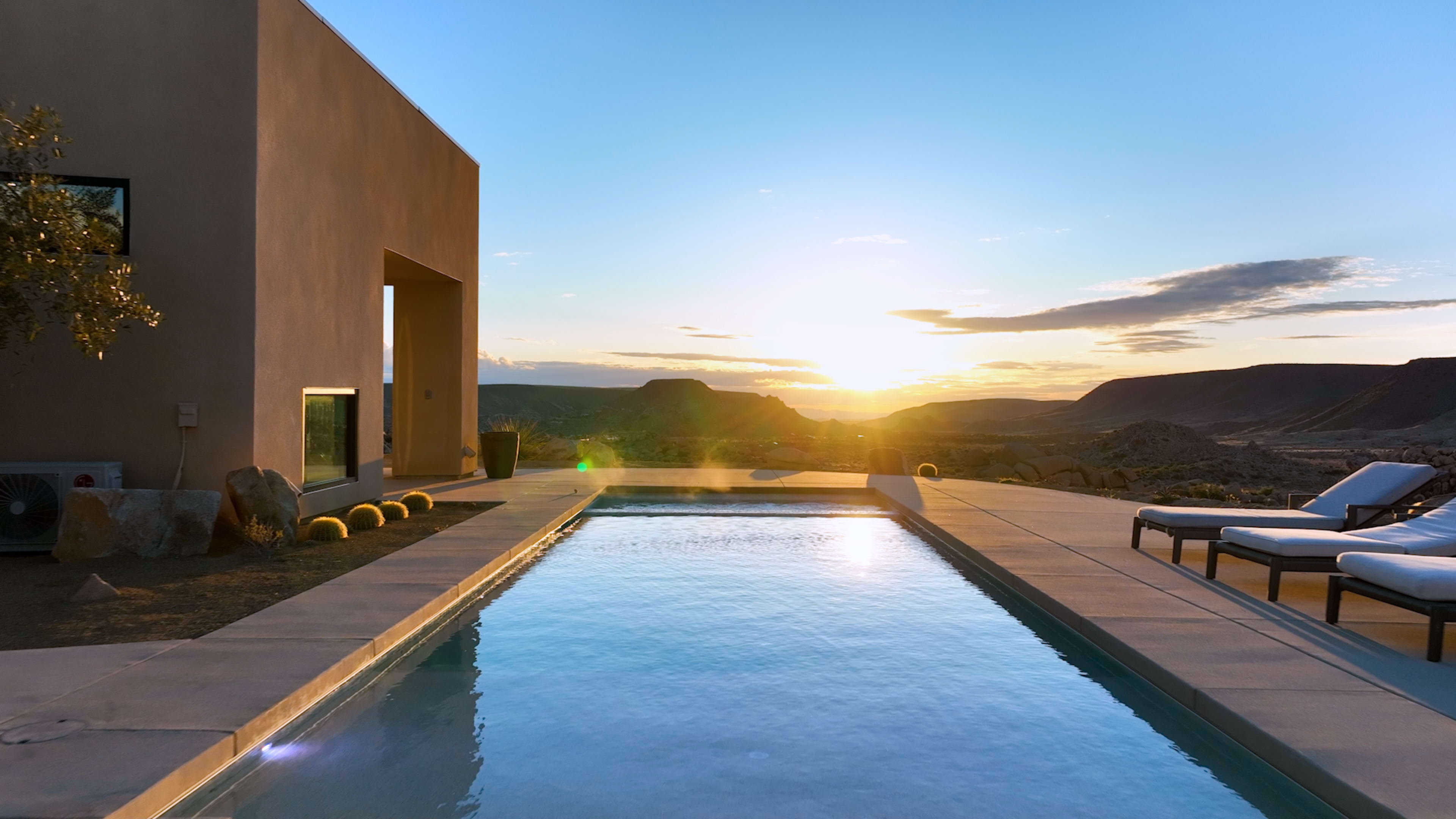 The image shows a modern pool area with lounge chairs, set against a backdrop of a sunset over a mountainous landscape.