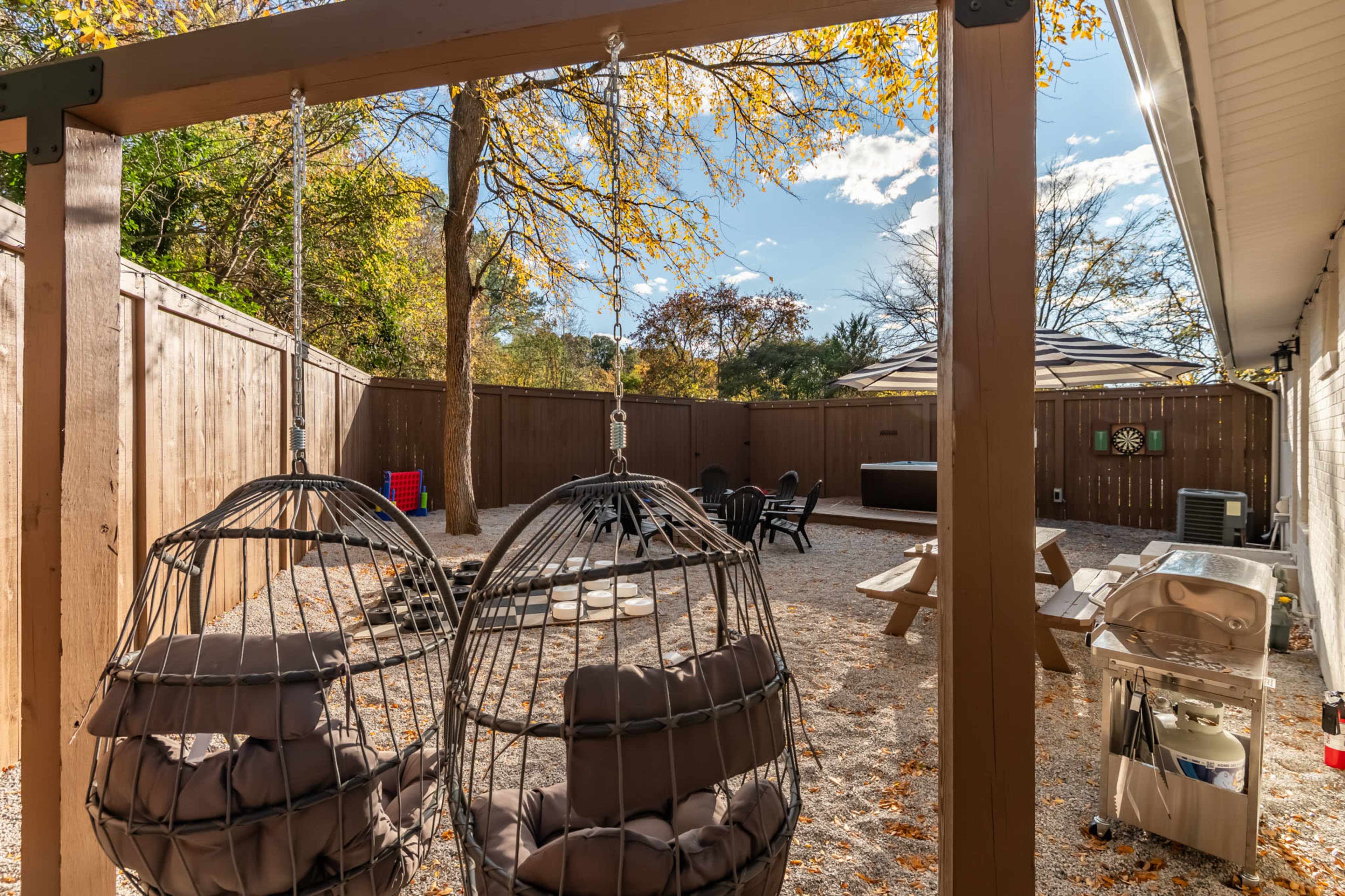 The image shows a backyard with two hanging chairs, a fire pit area surrounded by chairs, a picnic table, and a grill, all enclosed by wooden fencing and trees.