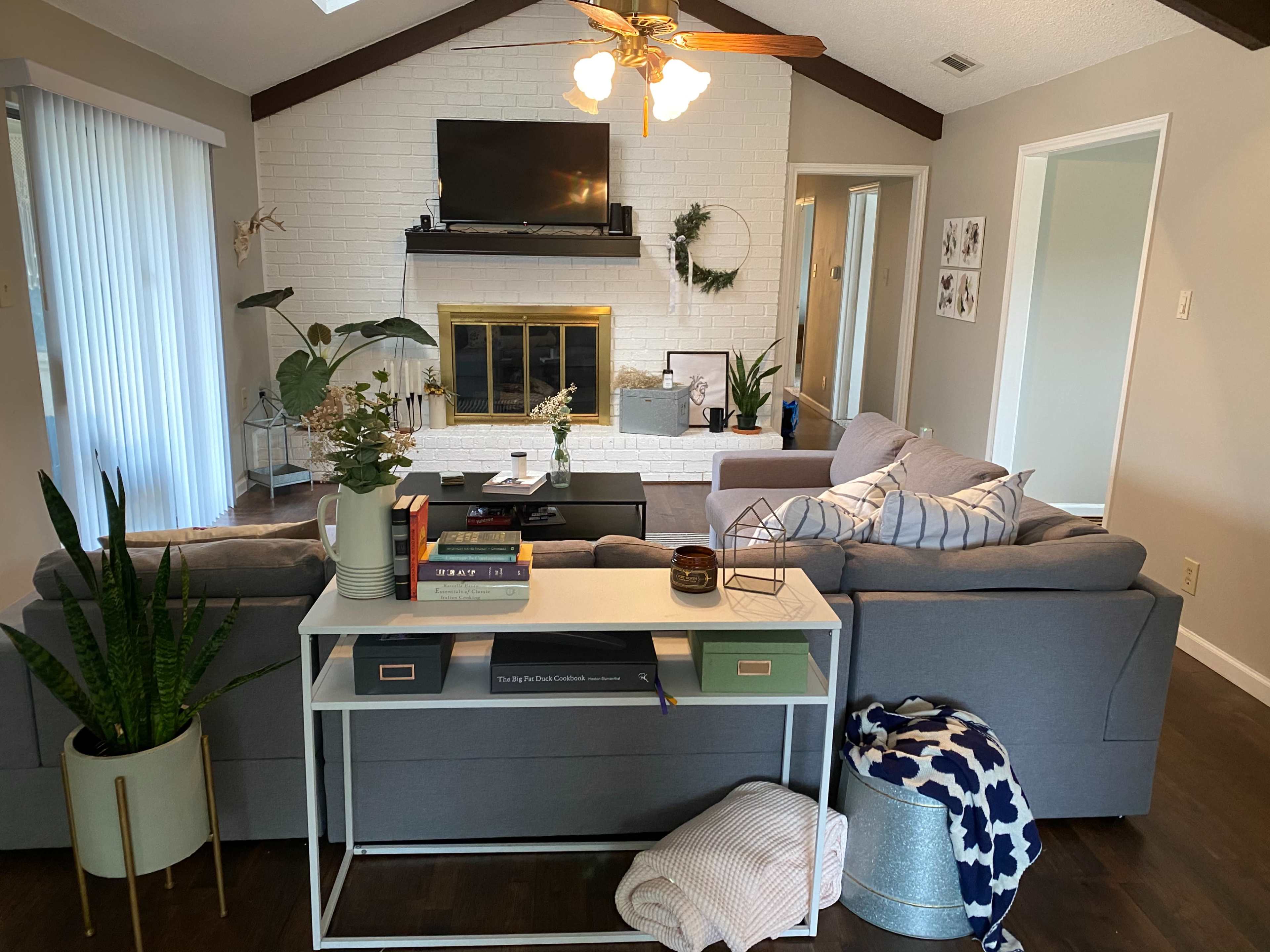 The living room features a gray sectional sofa facing a fireplace, with a coffee table in front and plants adorning the space.