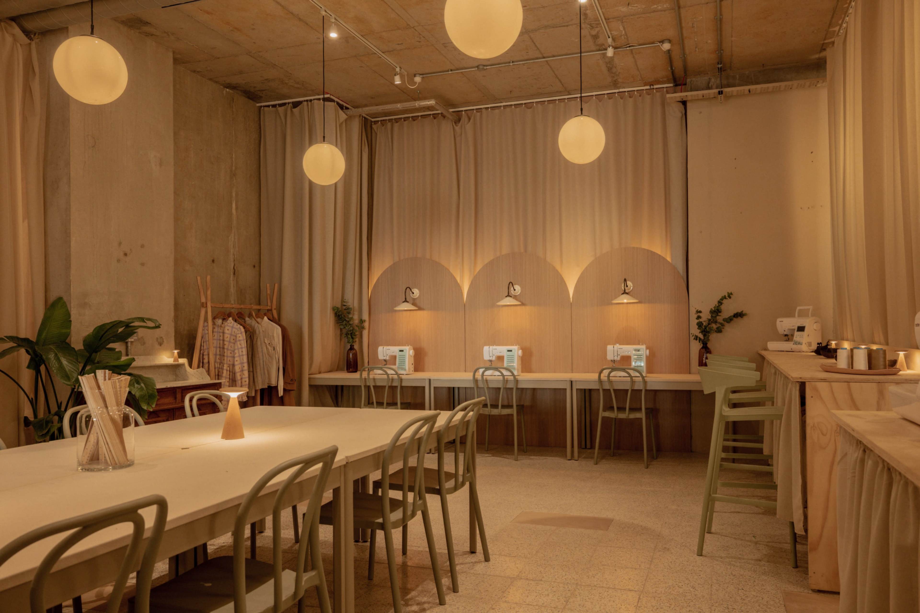 The image shows a neatly arranged sewing studio with several sewing machines, a long table, and soft lighting accentuated by round pendant lights.