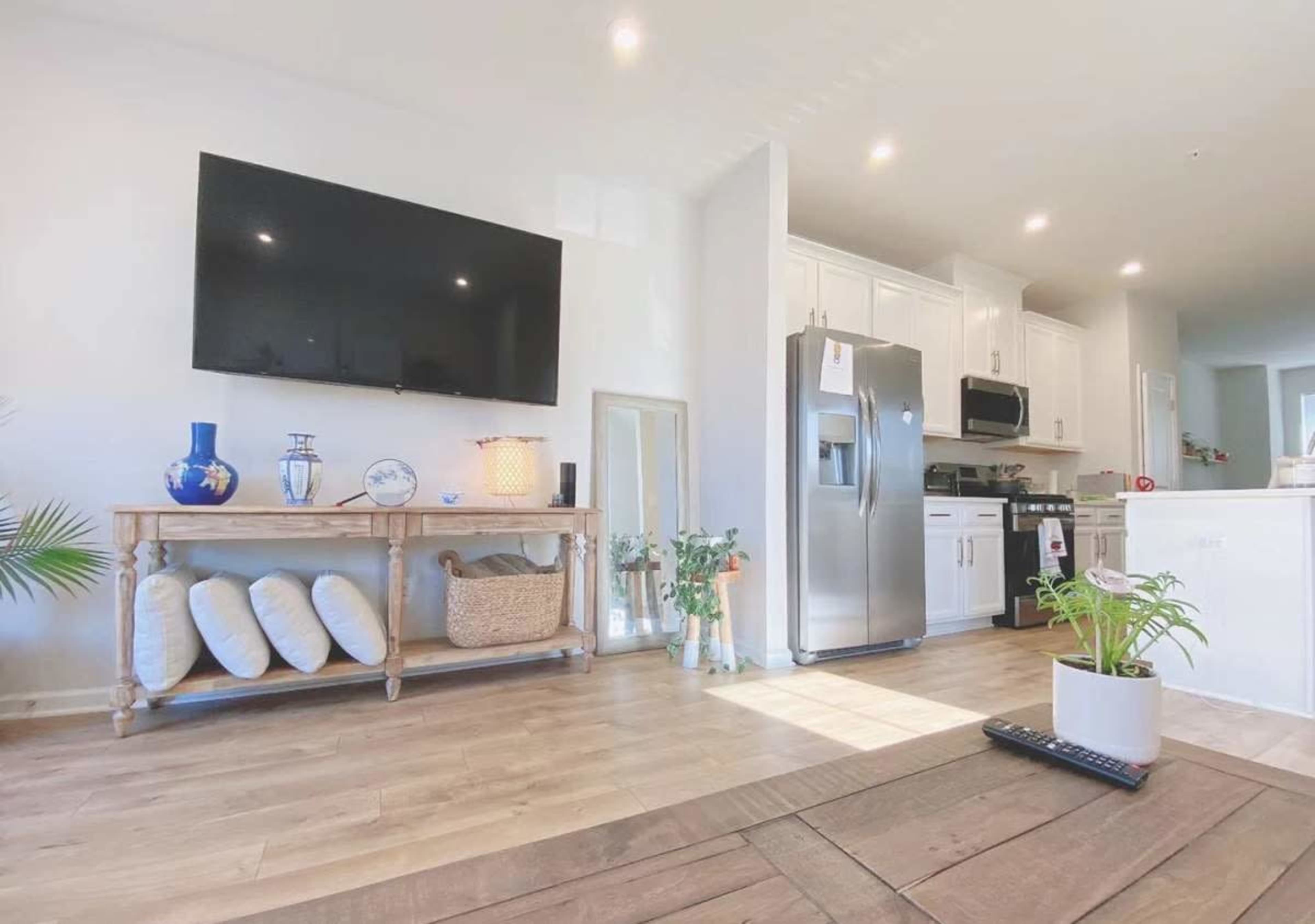 The image shows a modern kitchen and living area with a large flat-screen TV mounted on the wall, a wooden console table with decorative items, and a stainless steel refrigerator.