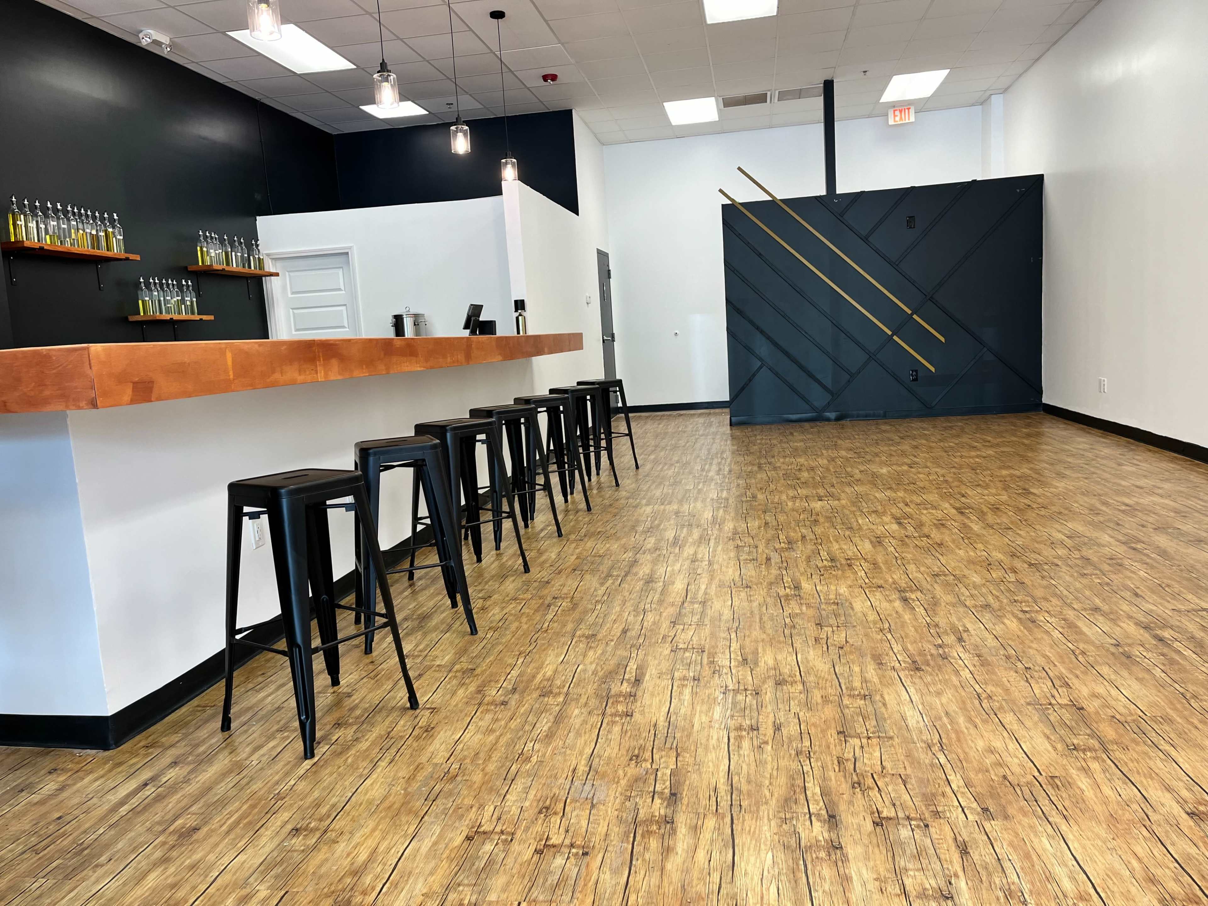 A modern, sparsely furnished interior space with a wooden bar, black metal stools, and a polished wooden floor.