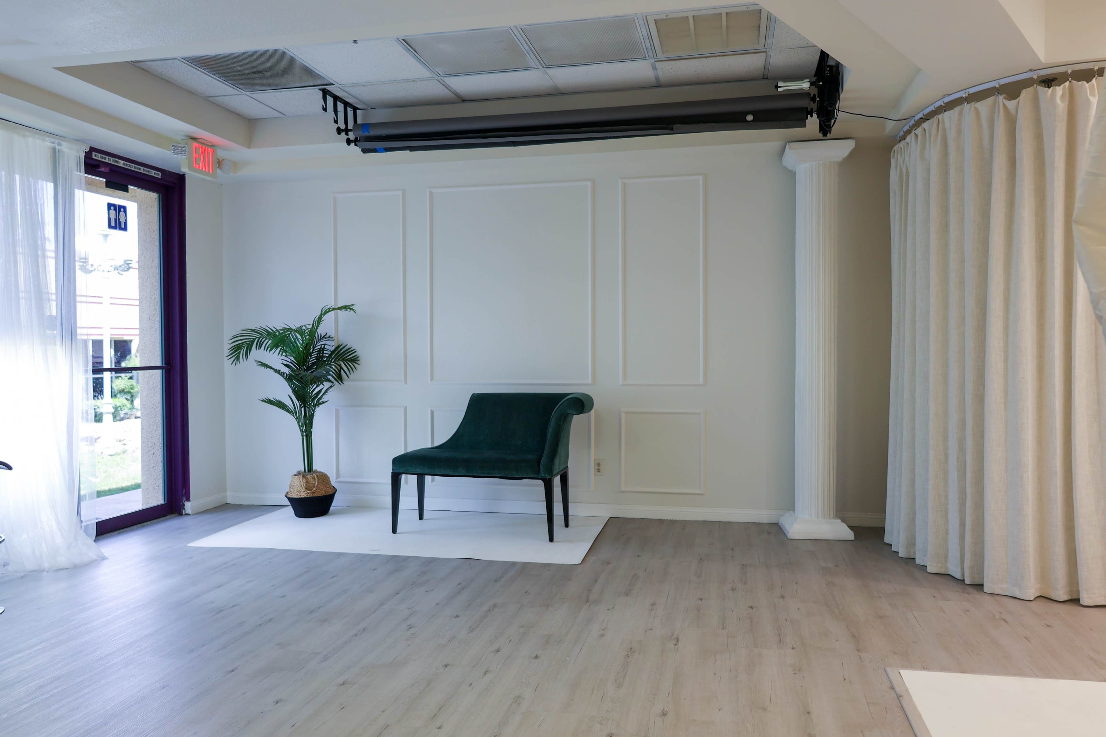 The image shows a minimalist indoor space with a green chair, a potted plant, and large windows, featuring light-colored walls and wooden flooring.