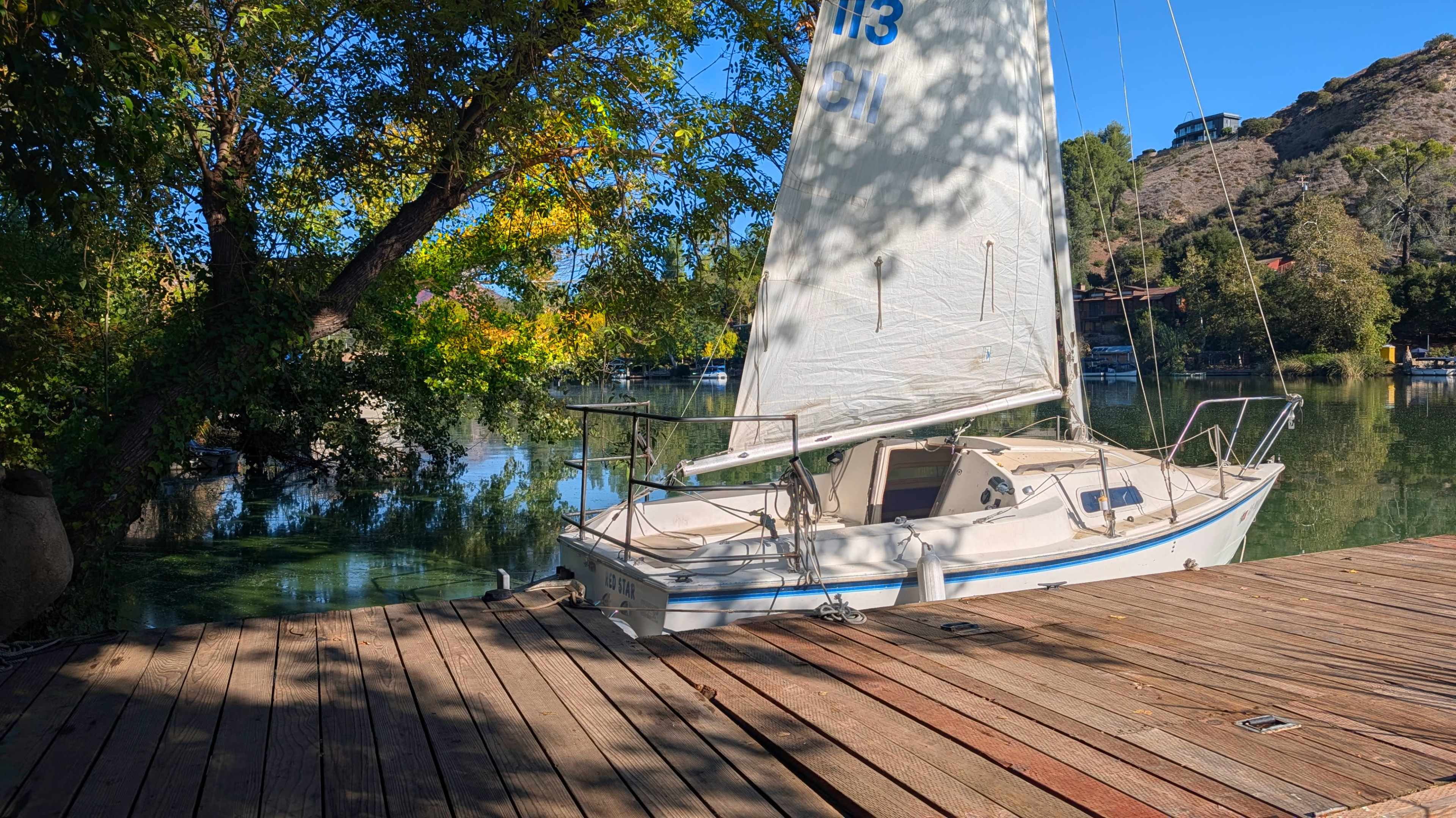 A small sailboat is docked on a wooden pier beside a calm body of water, surrounded by trees and hills.