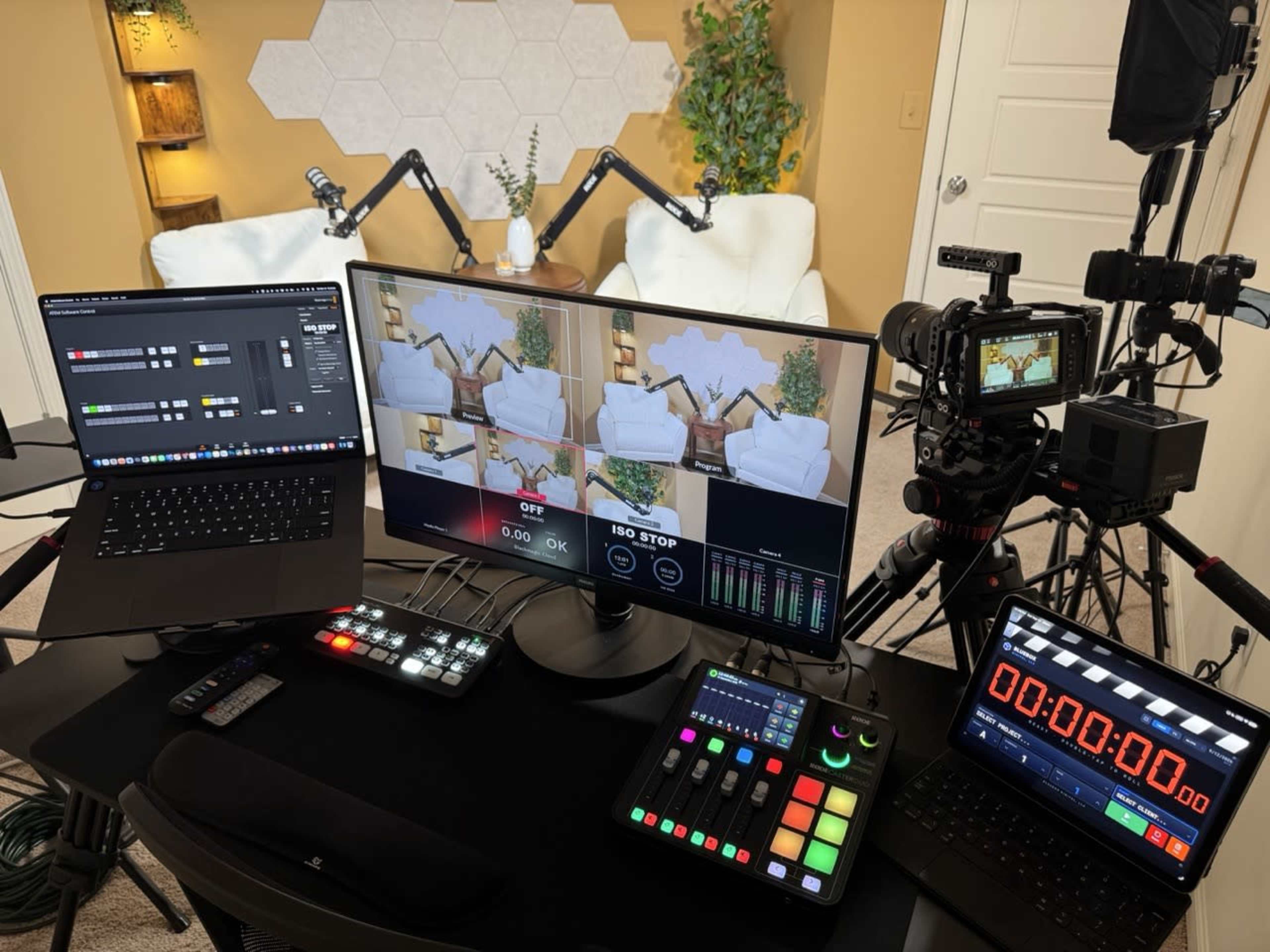 The image shows a media production setup featuring multiple screens displaying video feeds, audio equipment, and a camera positioned for recording in a well-lit room.