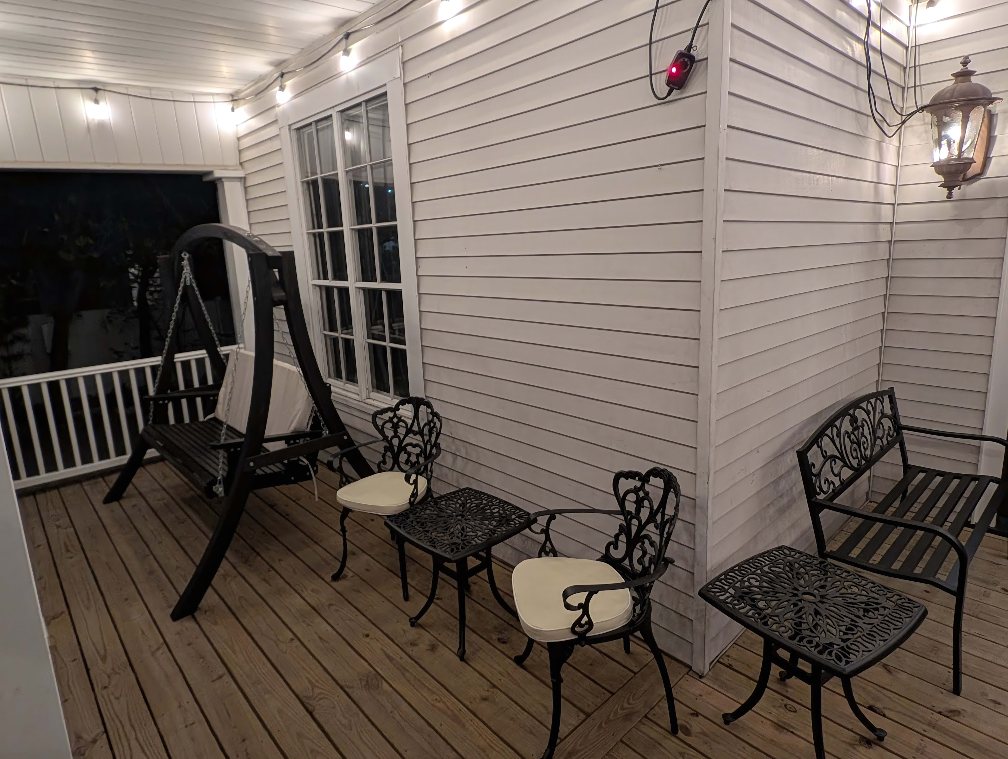The image shows a porch area with wooden flooring, multiple metal chairs, a swing, and decorative string lights overhead.
