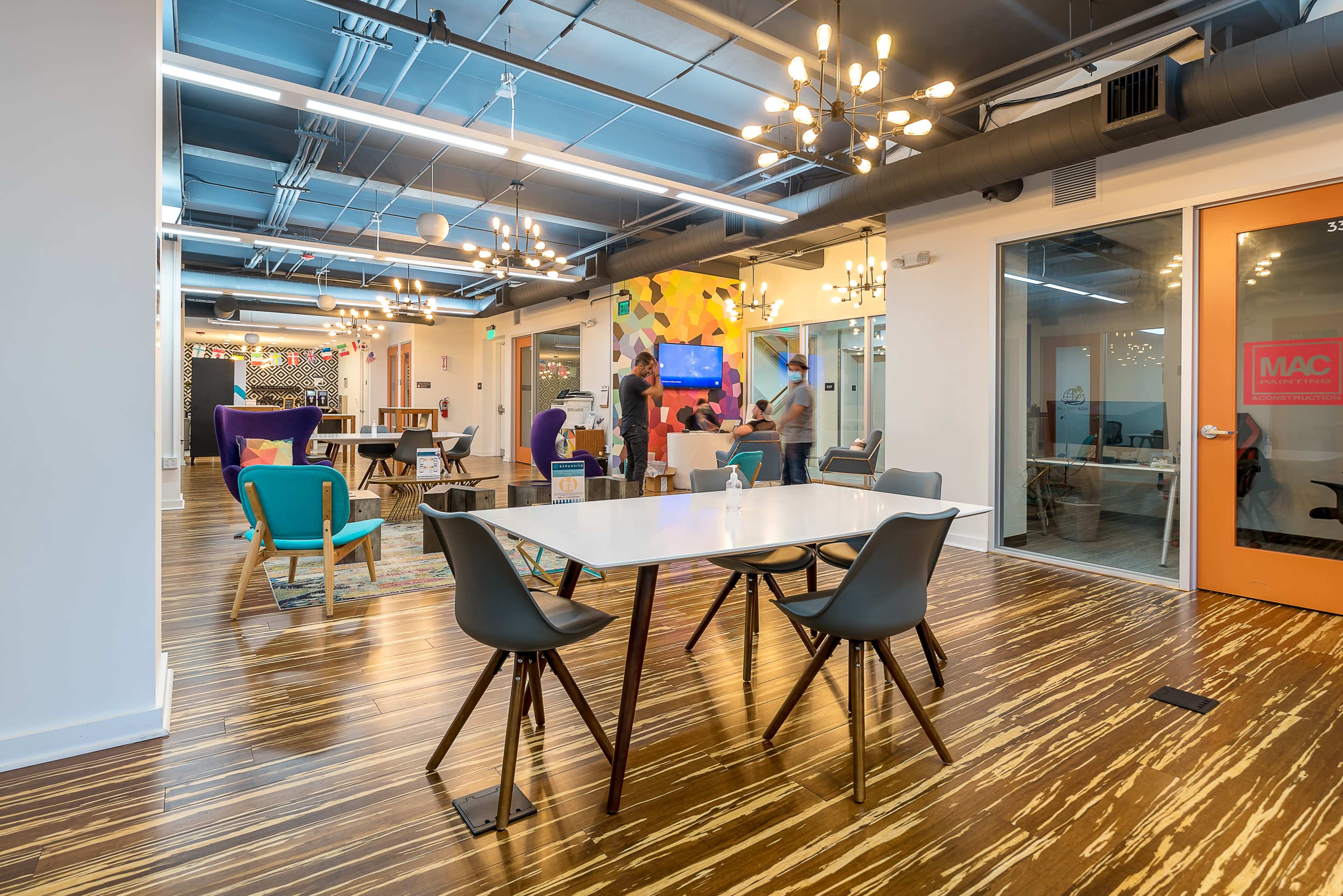The image shows a modern office space featuring a central table surrounded by chairs, decorative lighting fixtures, and colorful wall art, with people engaged in activities across different areas.