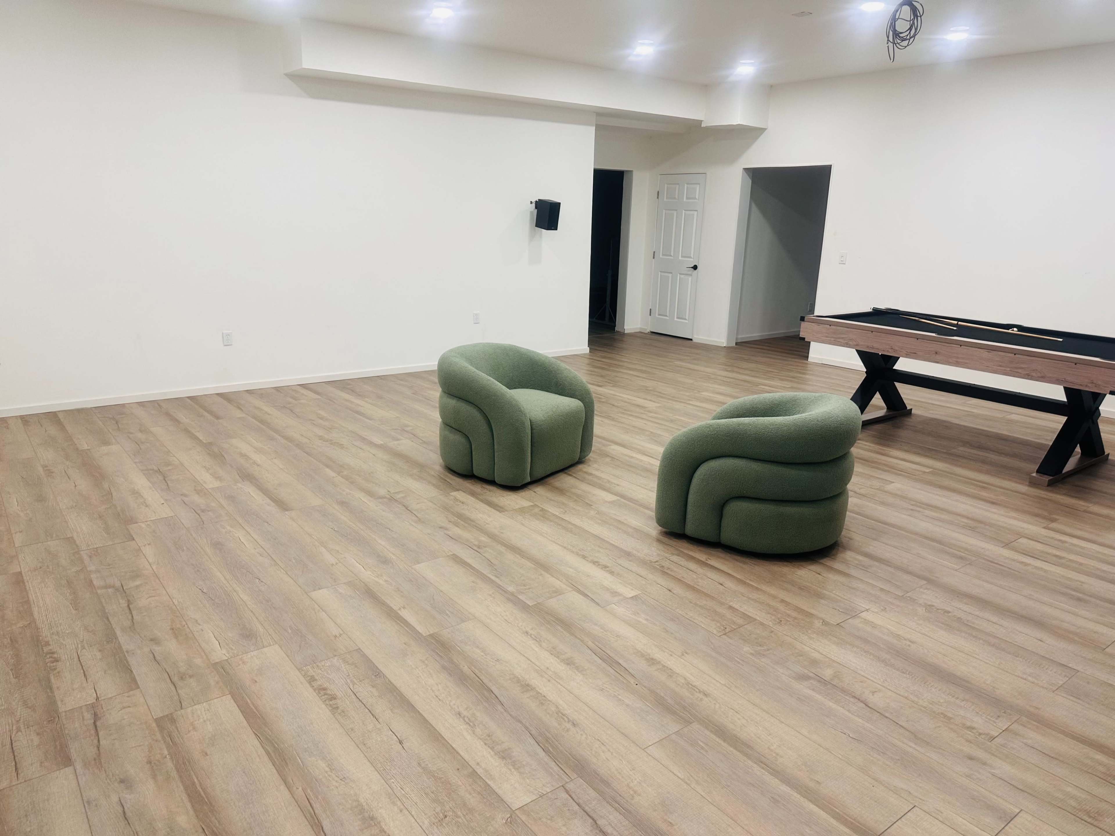 The image shows a spacious room with light wooden flooring, featuring two green armchairs and a pool table in the background.