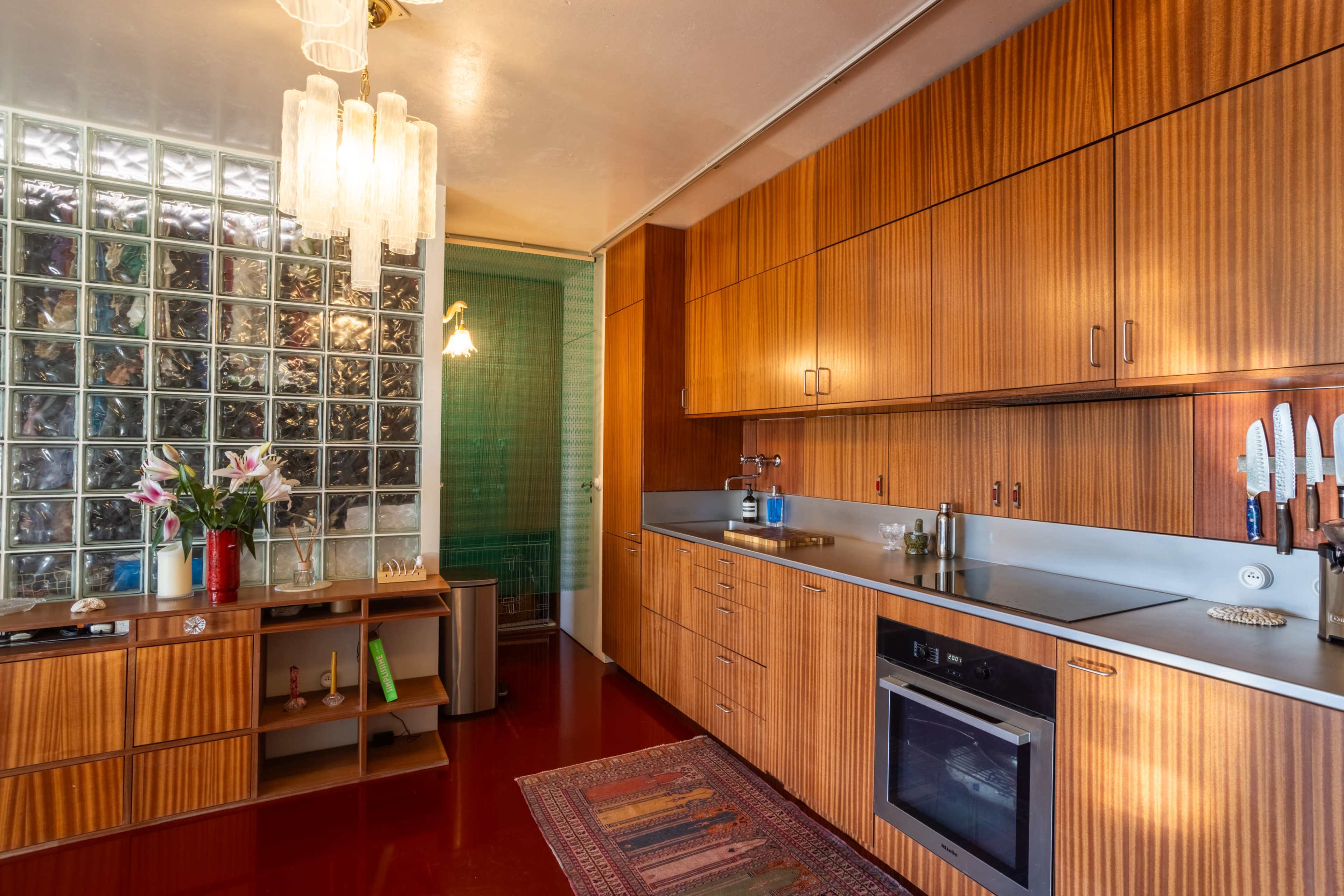 The image shows a kitchen featuring wooden cabinetry, a glass block wall, and a small area with a decorative rug.