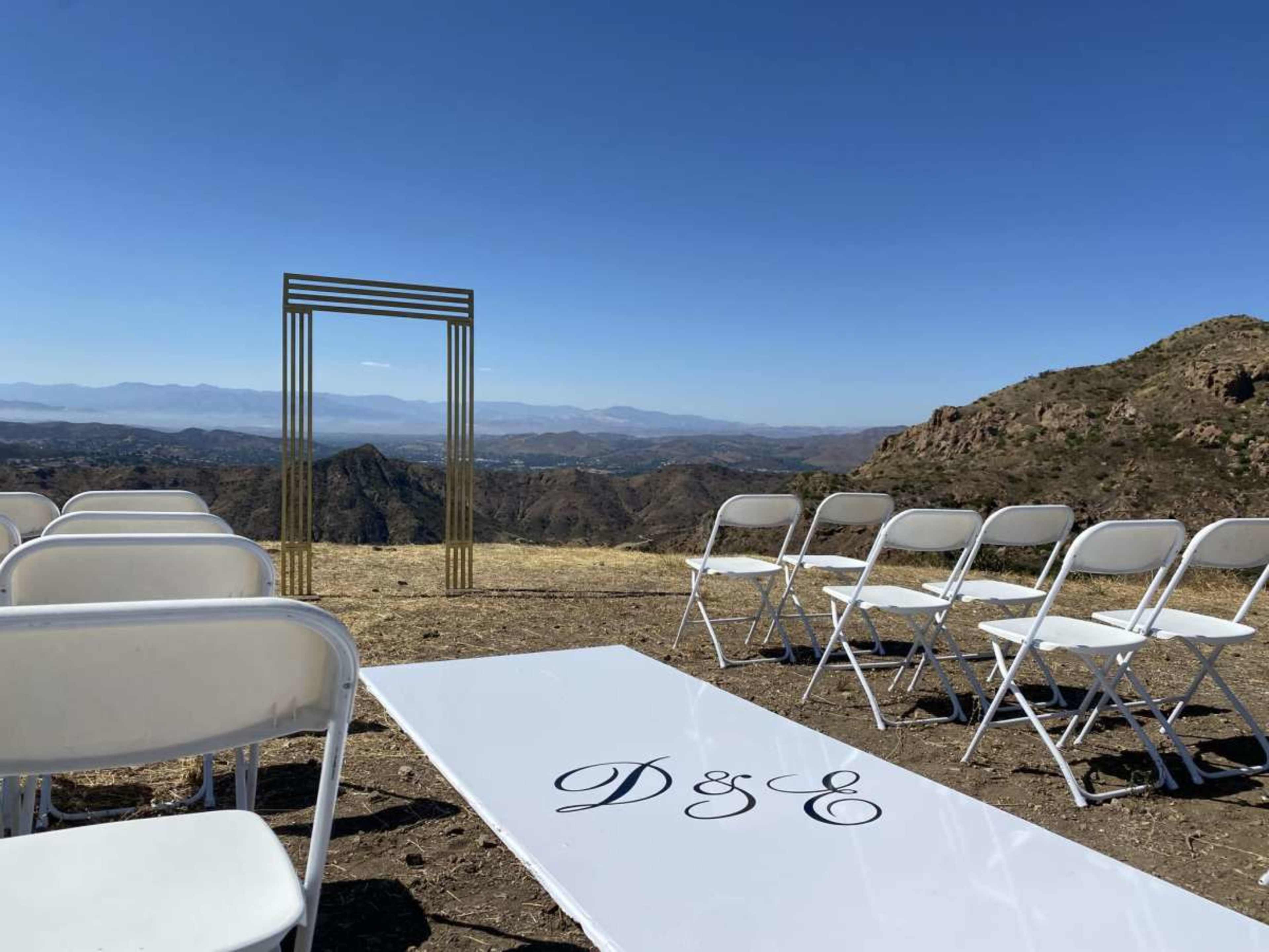 A wedding setup with white chairs and a decorated aisle leading to a gold arch overlooking a mountainous landscape.