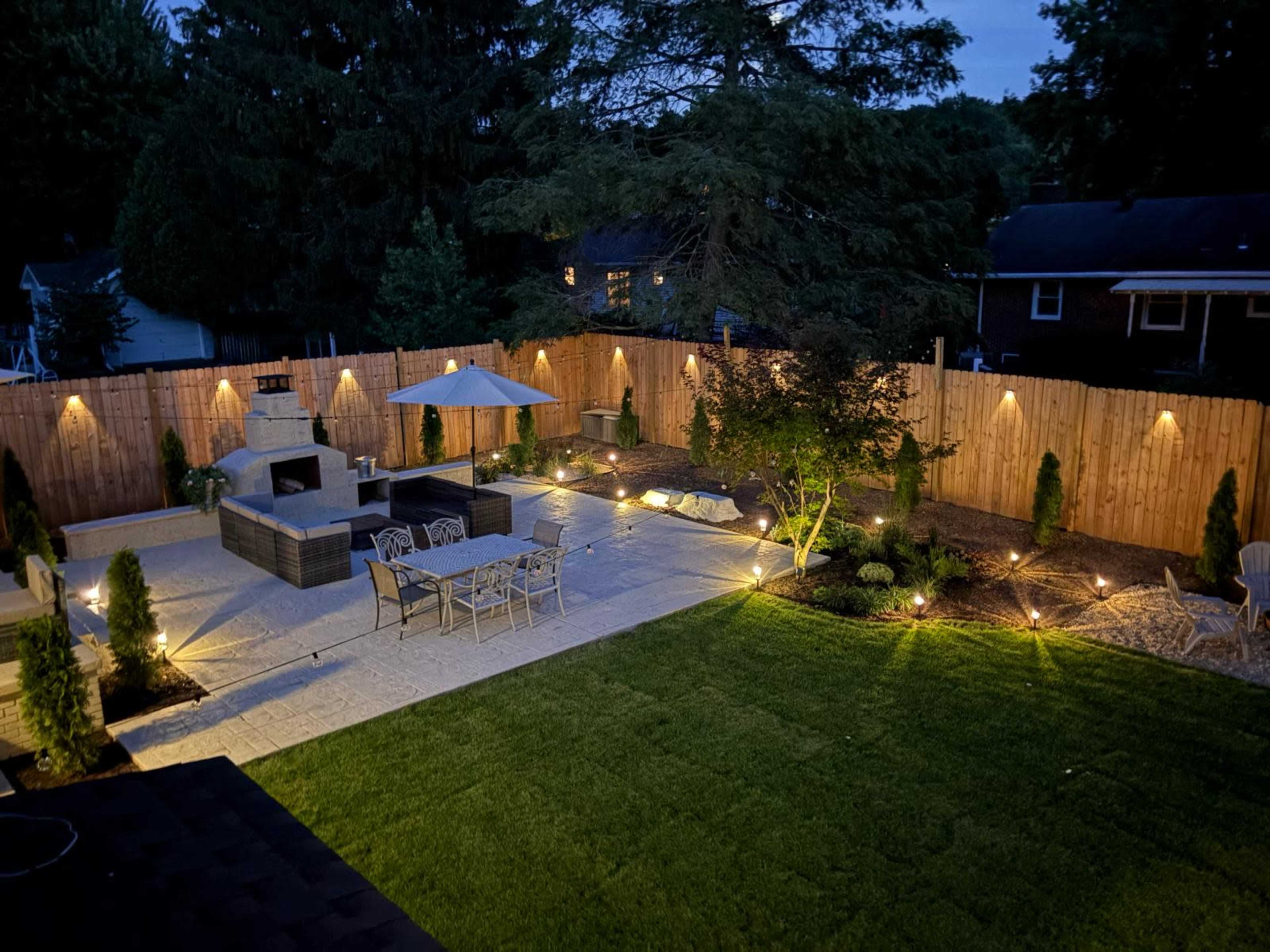 A well-lit backyard patio features a seating area, a dining table, and landscaping with trees and shrubs, all surrounded by a wooden fence.