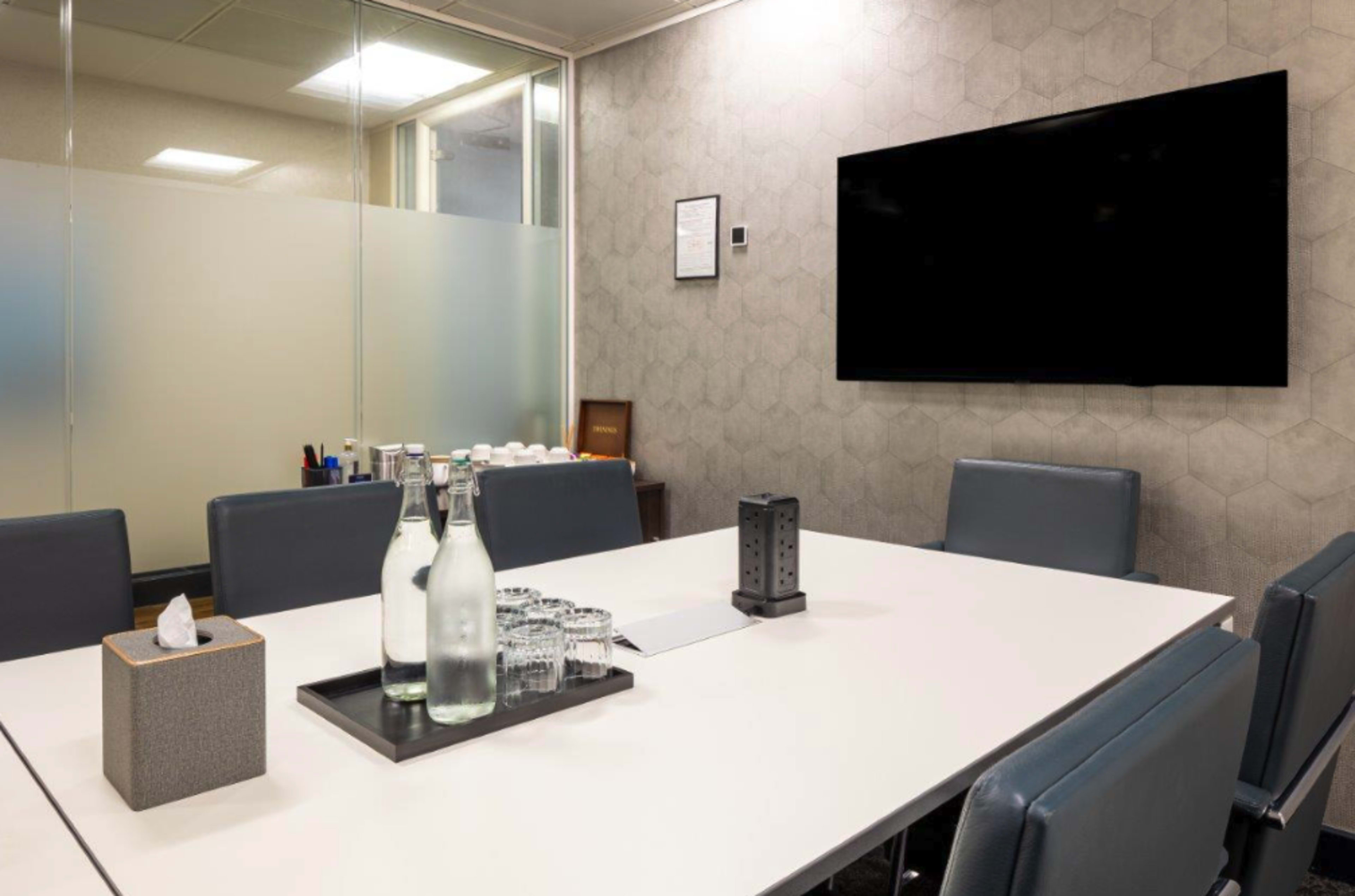 A glass-walled meeting room features a rectangular table set with water bottles, glasses, and a notepad, alongside a wall-mounted television and a geometric-patterned wallpaper.