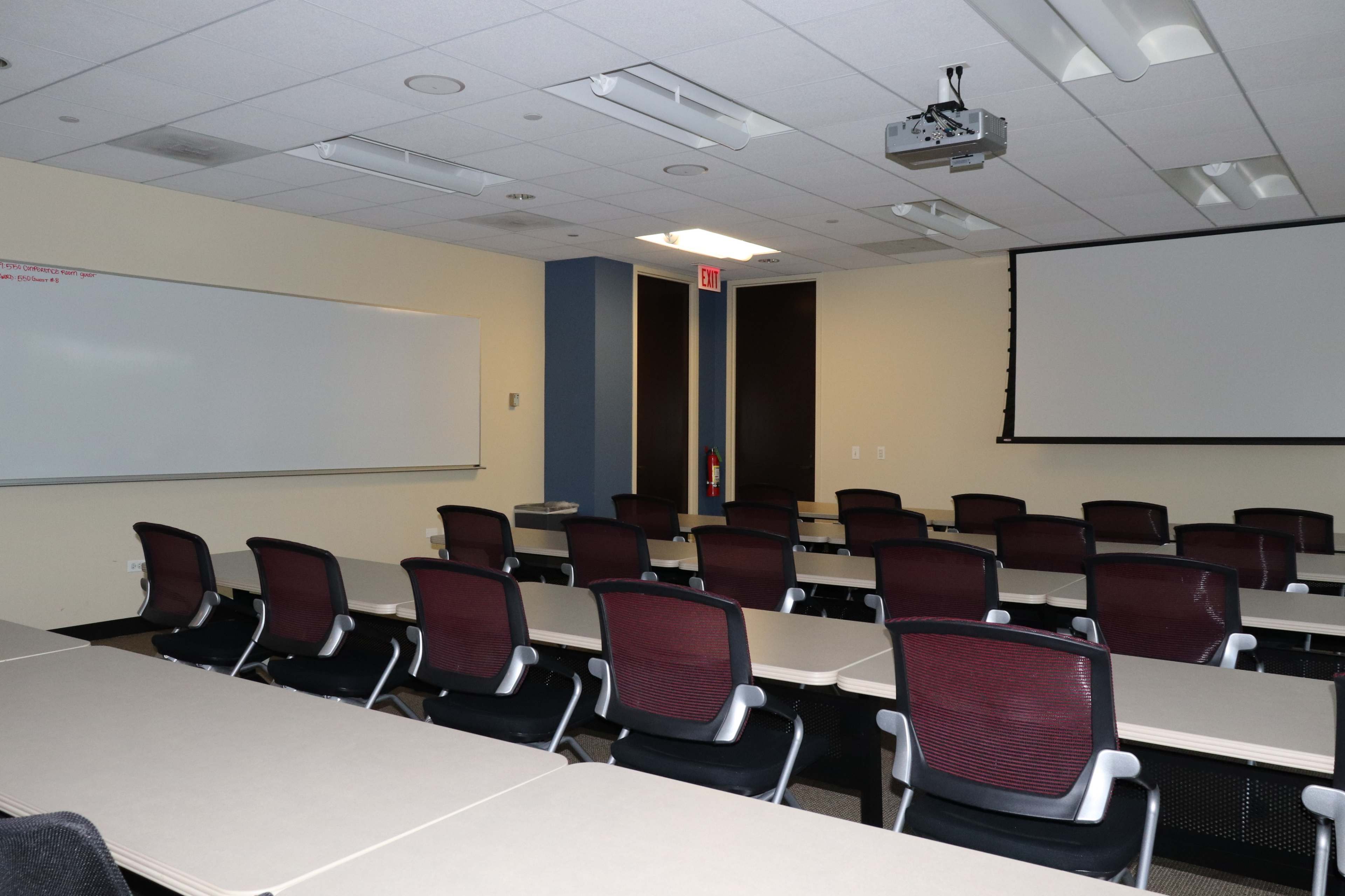 A classroom with rows of tables and chairs, a whiteboard on one wall, and a projector mounted on the ceiling.