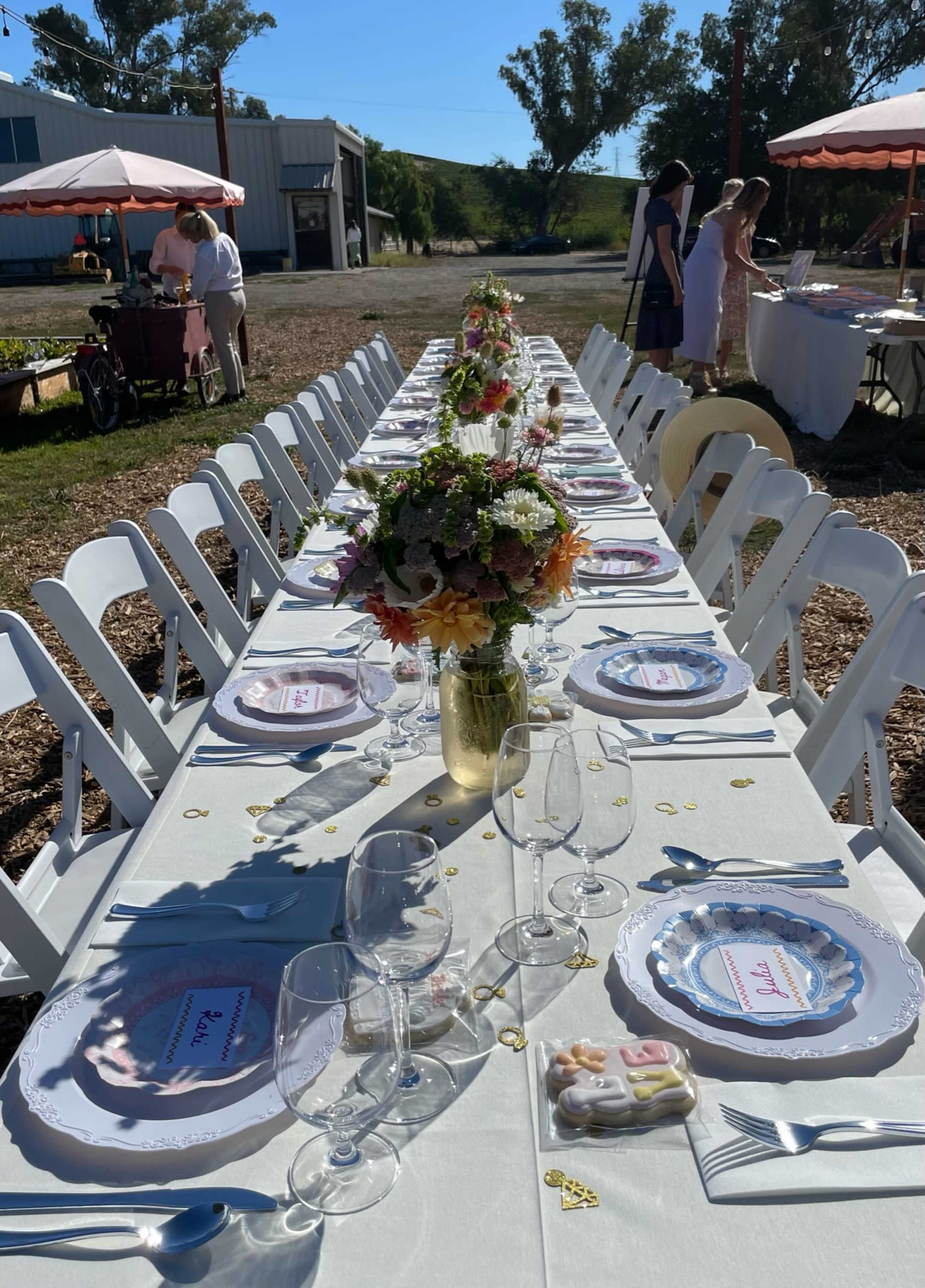 A long outdoor dining table is set with plates, glasses, and floral centerpieces under a clear blue sky.
