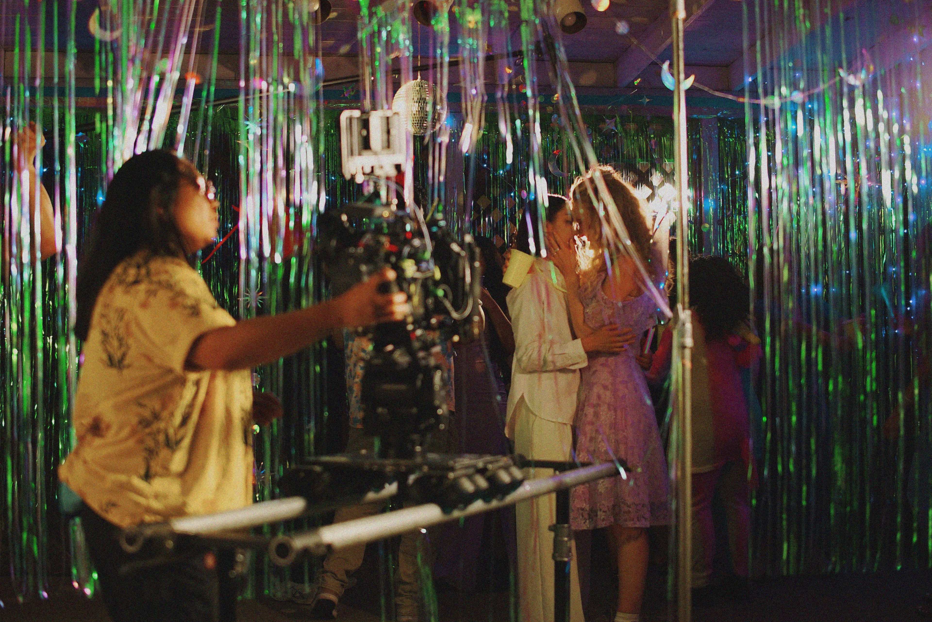 A film crew captures a romantic scene between two actors in a festively decorated setting with shimmering streamers.