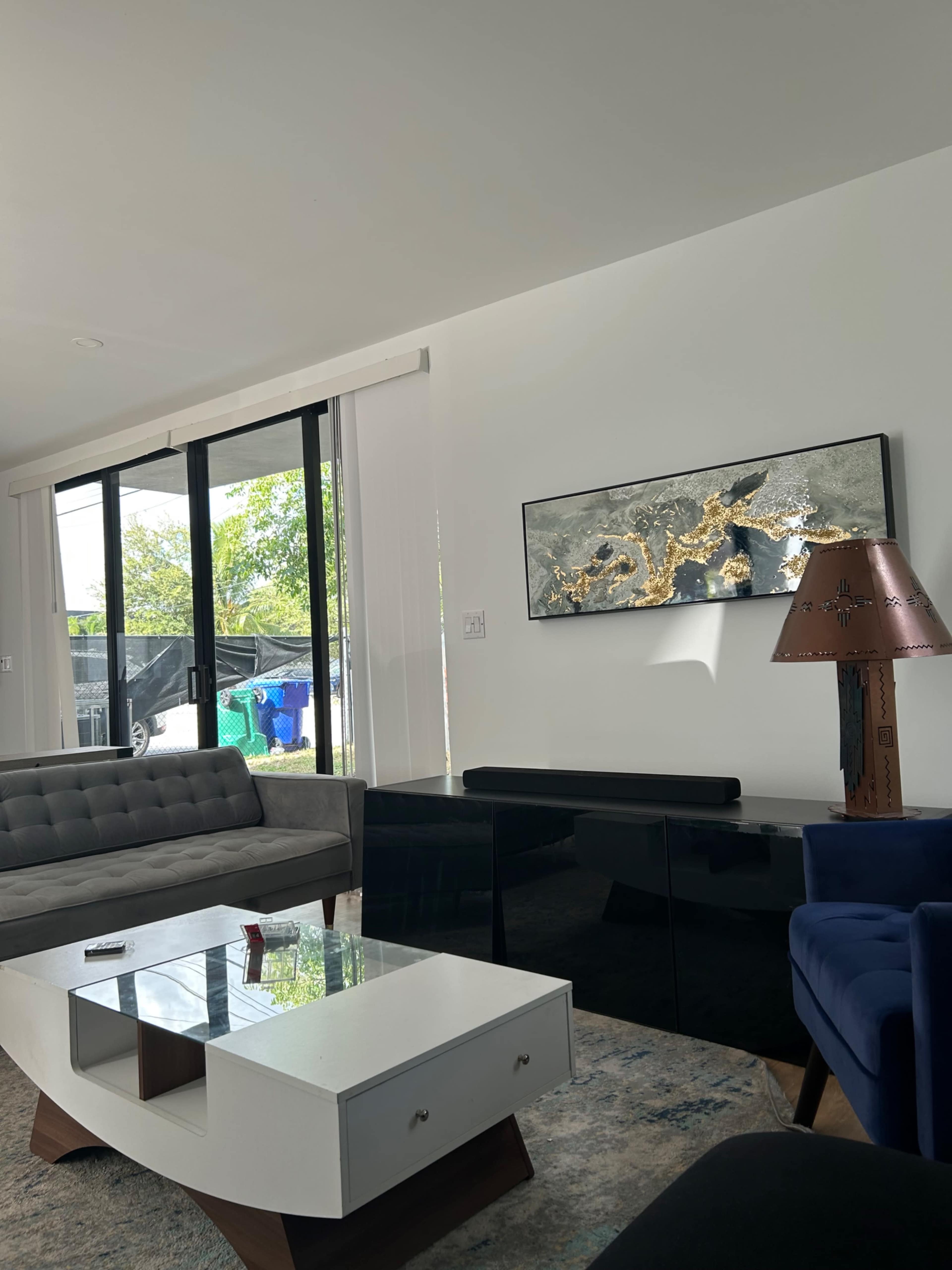The image shows a modern living room with a gray sofa, a blue armchair, a white coffee table, large windows allowing natural light, and a decorative wall art piece.