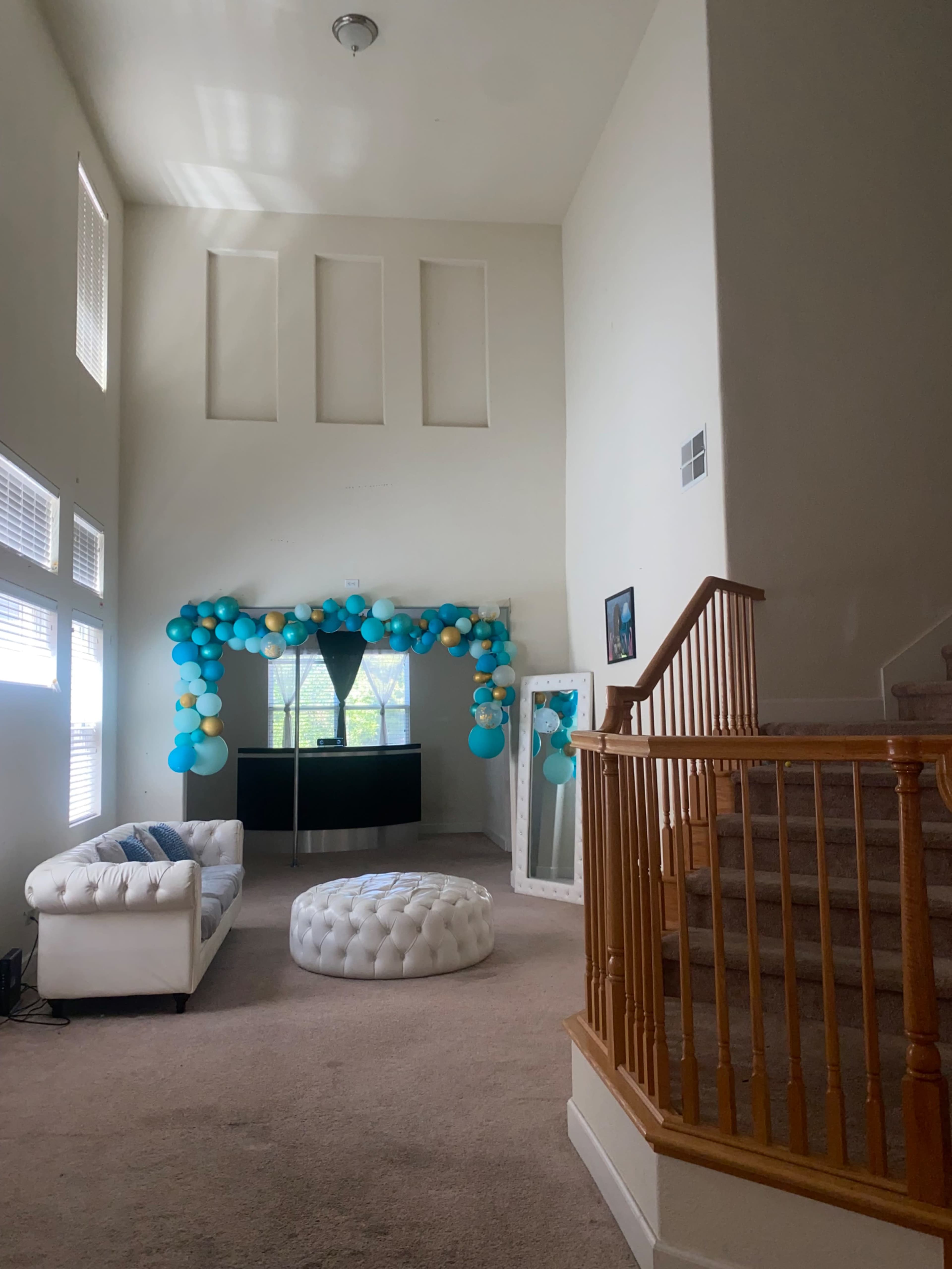 A spacious living room with a white couch and a tufted ottoman, decorated with a balloon arch in blue and gold, and a staircase leading to an upper level.