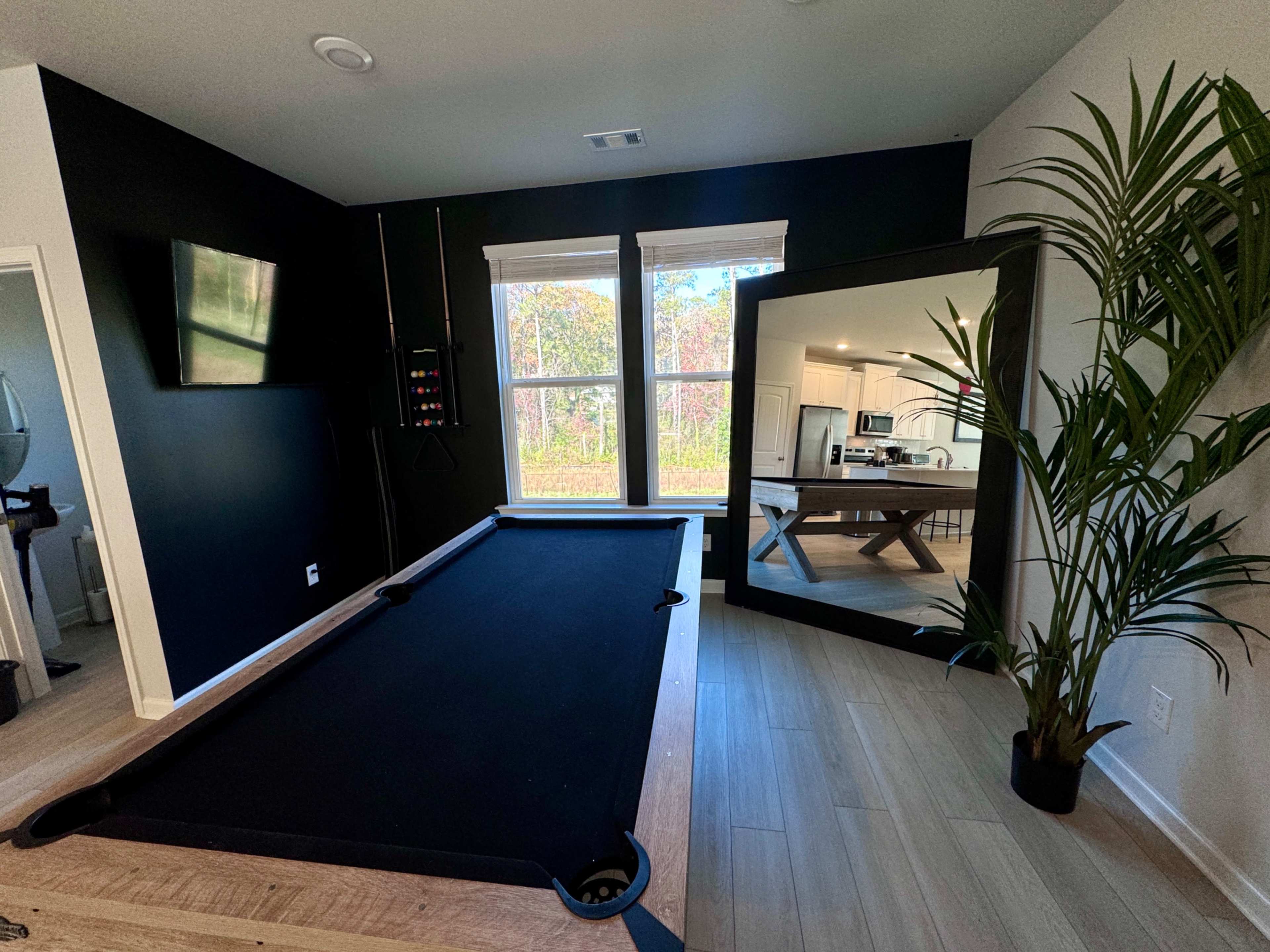 The image shows a room with a pool table on a wooden floor, large windows providing natural light, a mirror, and a potted plant.