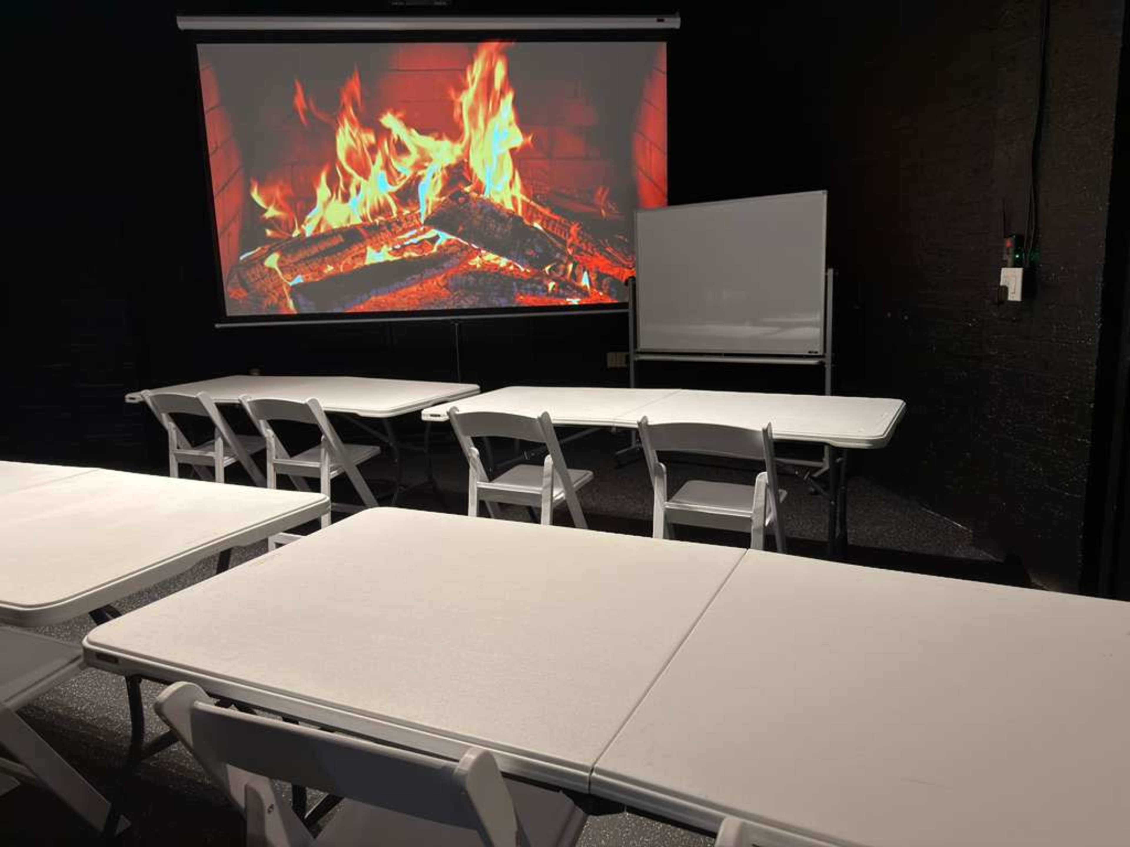 A room set up for a meeting features white folding tables arranged in rows, facing a large screen displaying a crackling fire.