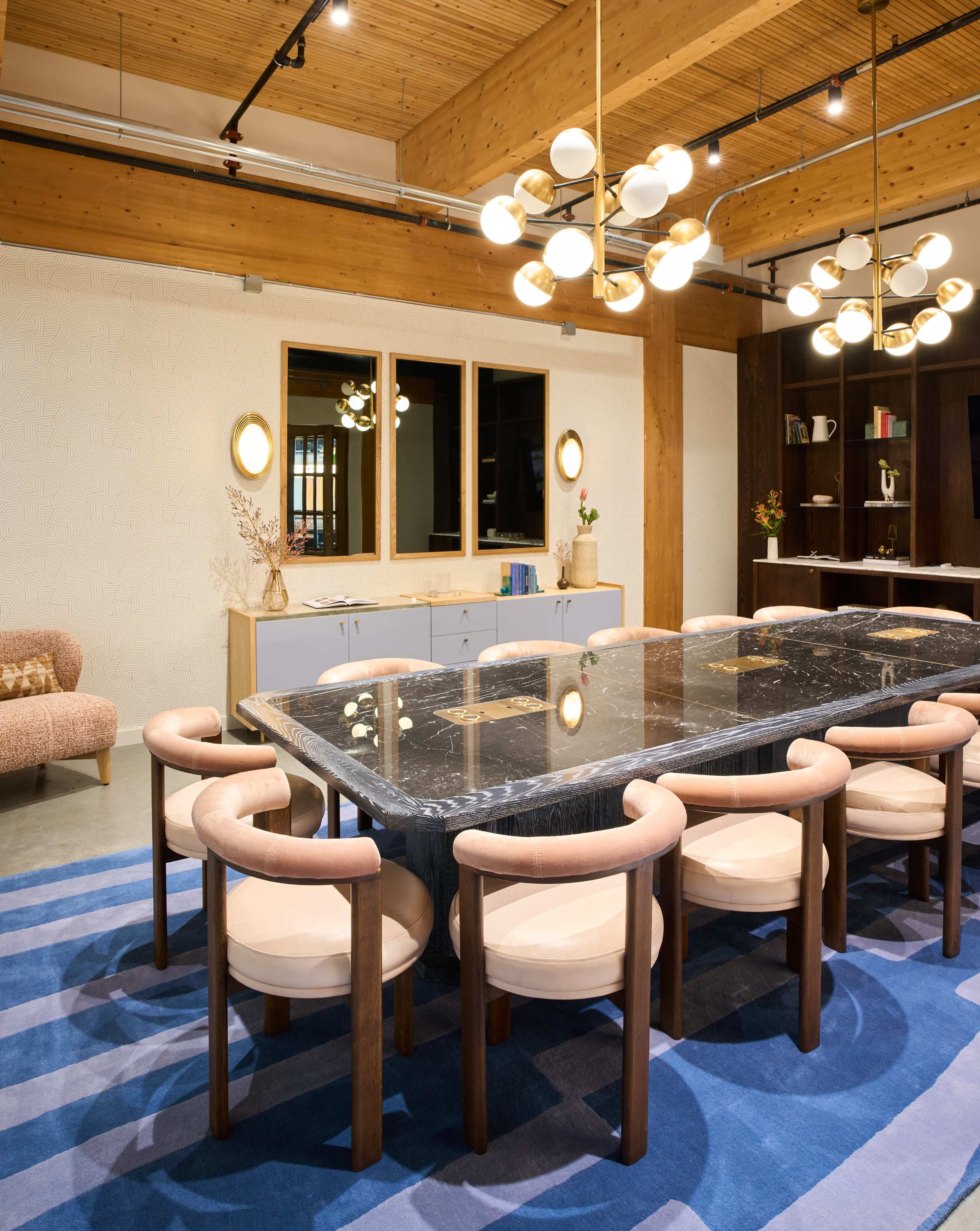 The image shows a modern dining area with a large black marble table surrounded by light-colored chairs, complemented by a patterned rug and contemporary lighting fixtures.