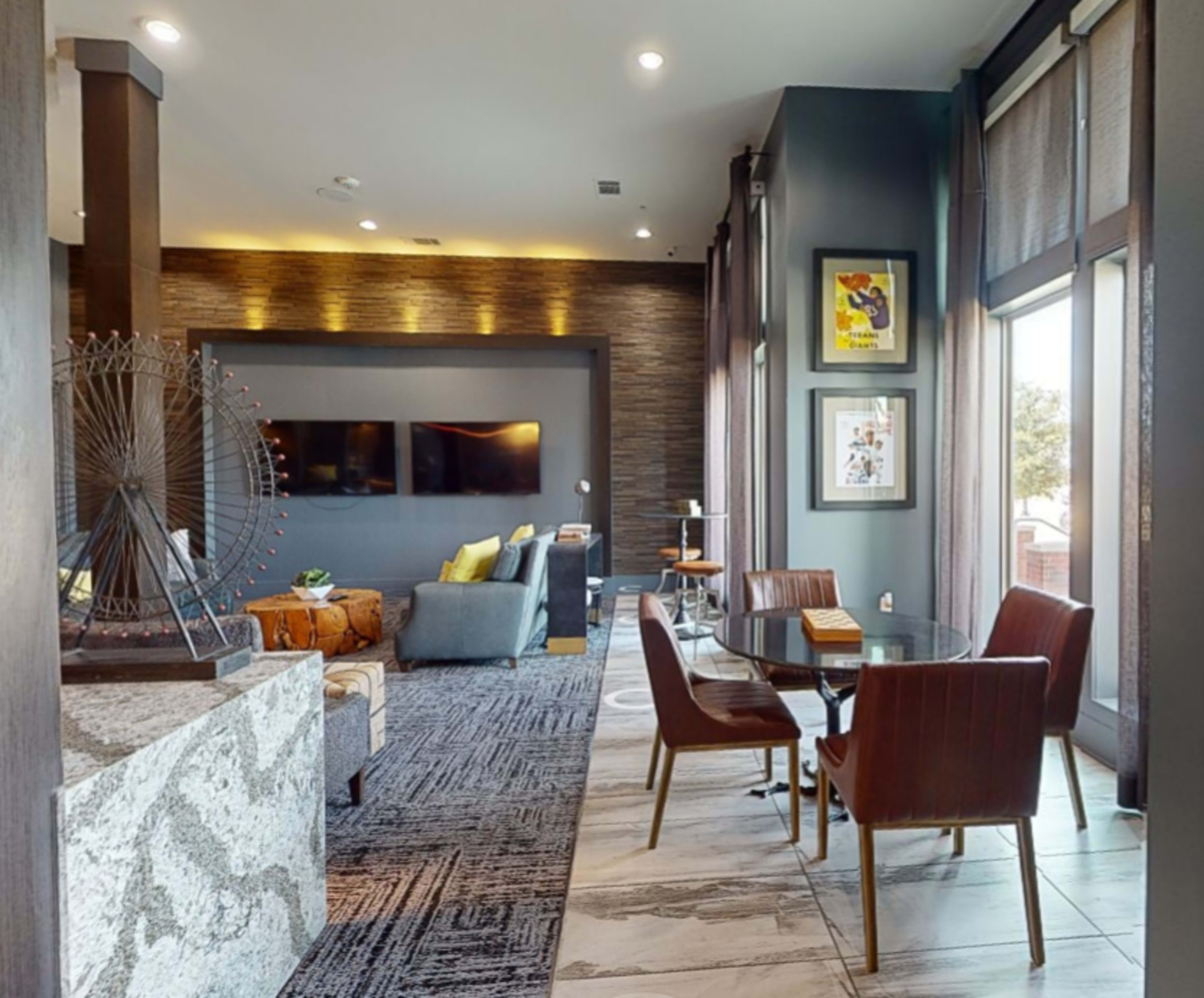 A modern living space featuring a seating area with a large television, a dining table, and large windows allowing natural light.