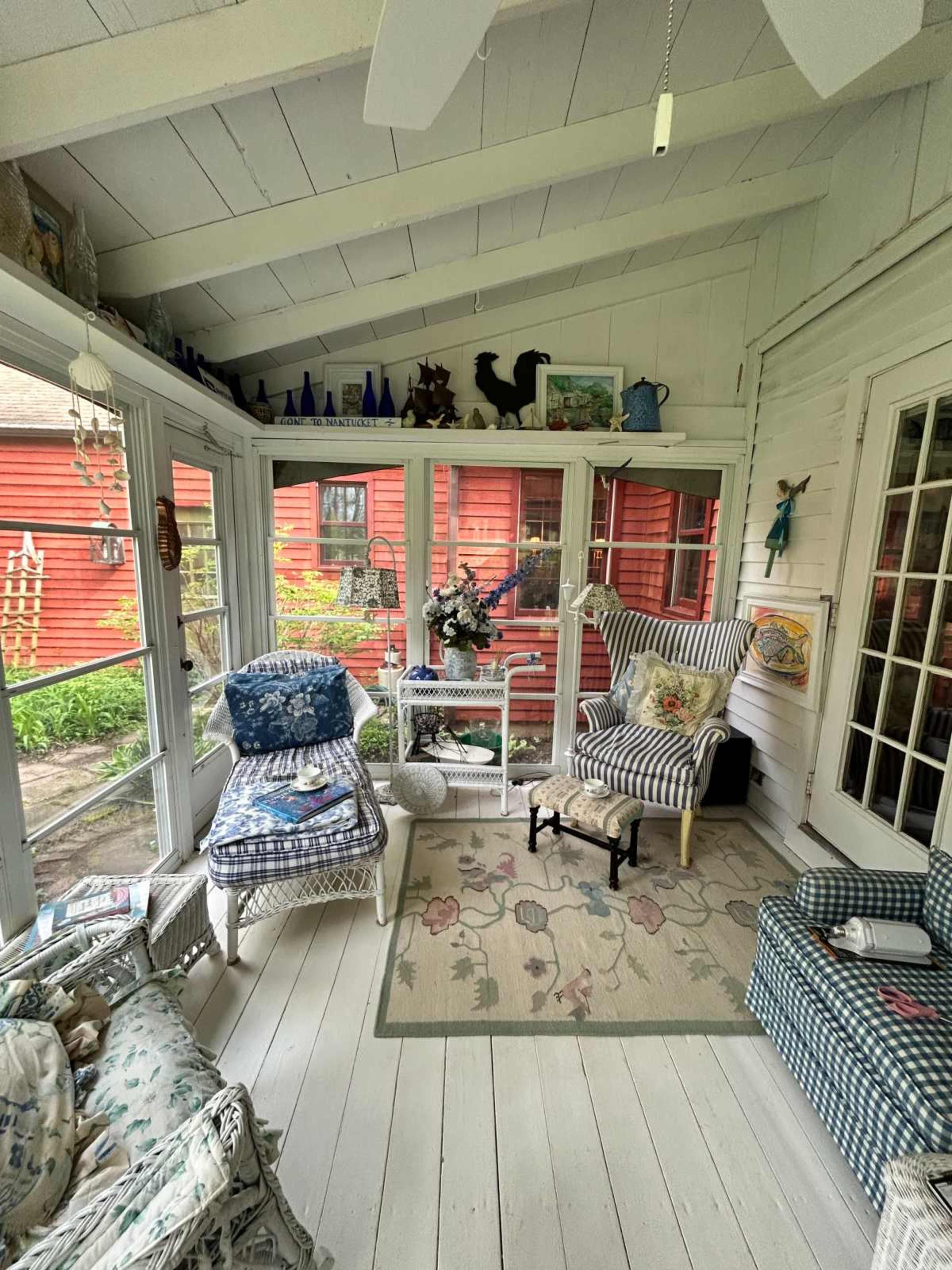 The image shows a cozy sunroom with white wooden walls, various patterned furniture, and a view of a red building outside through the glass panels.