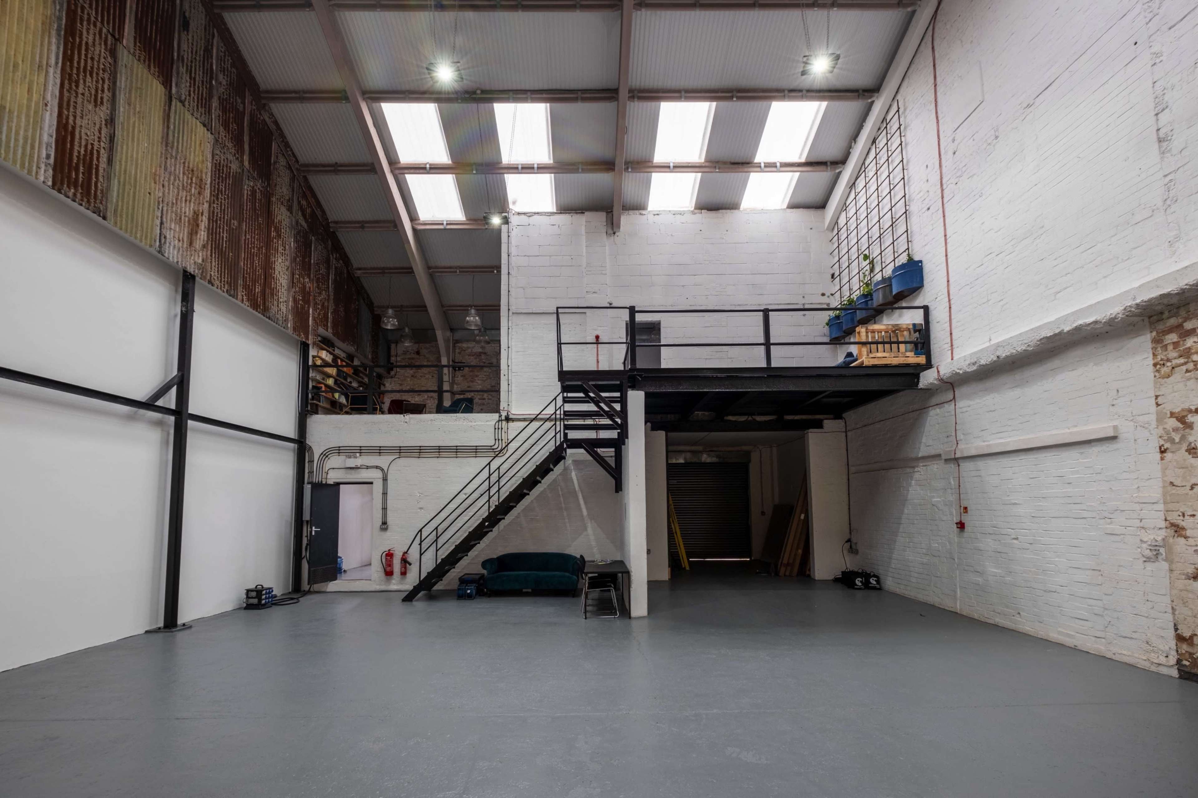 An empty warehouse interior featuring a mezzanine level with a staircase, exposed brick walls, and large windows.