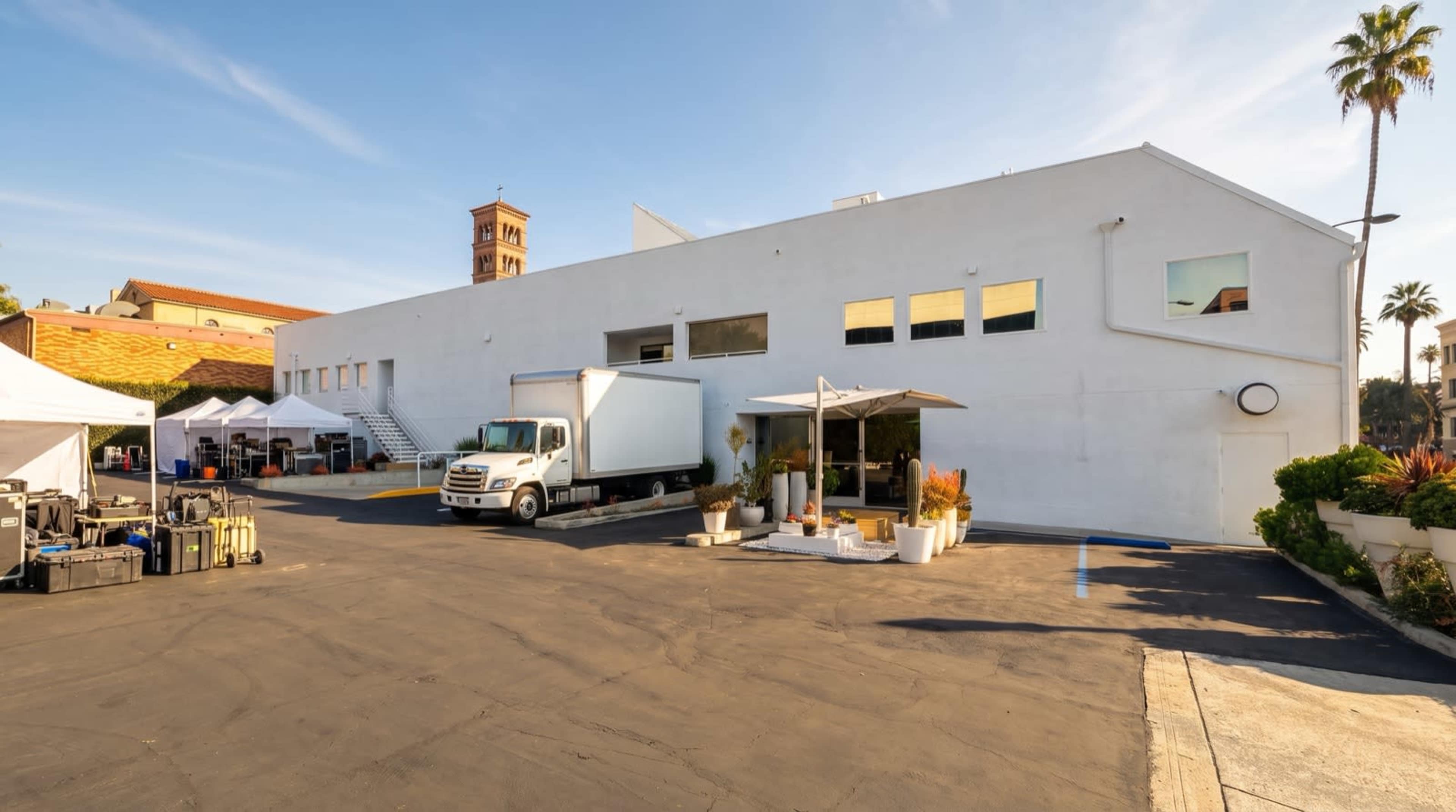A white commercial building with a loading area and a parked delivery truck is situated in a parking lot, surrounded by palm trees and outdoor tents.
