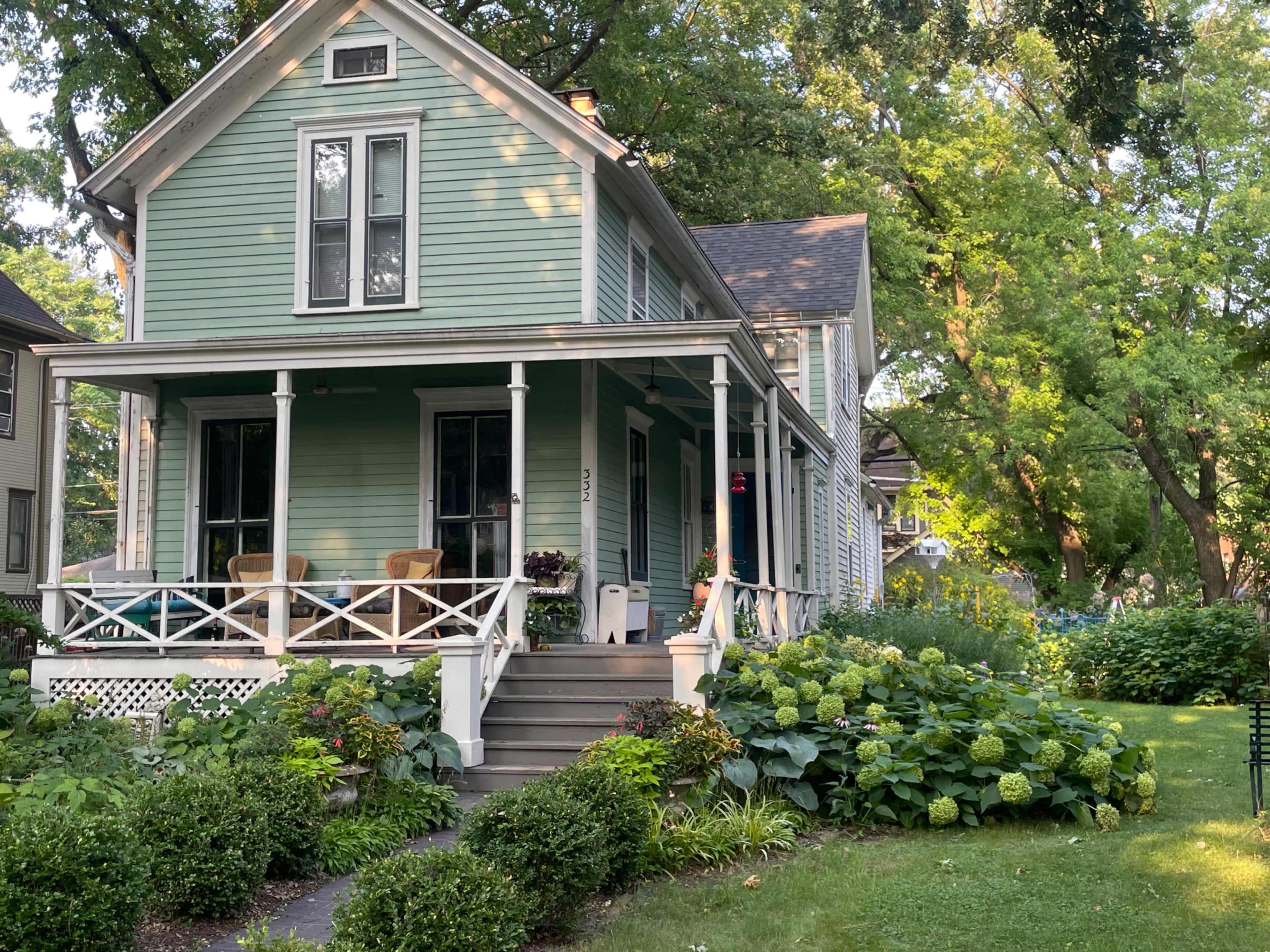 A green two-story house with afront porch and flower-filled garden is surrounded by trees and shrubs.