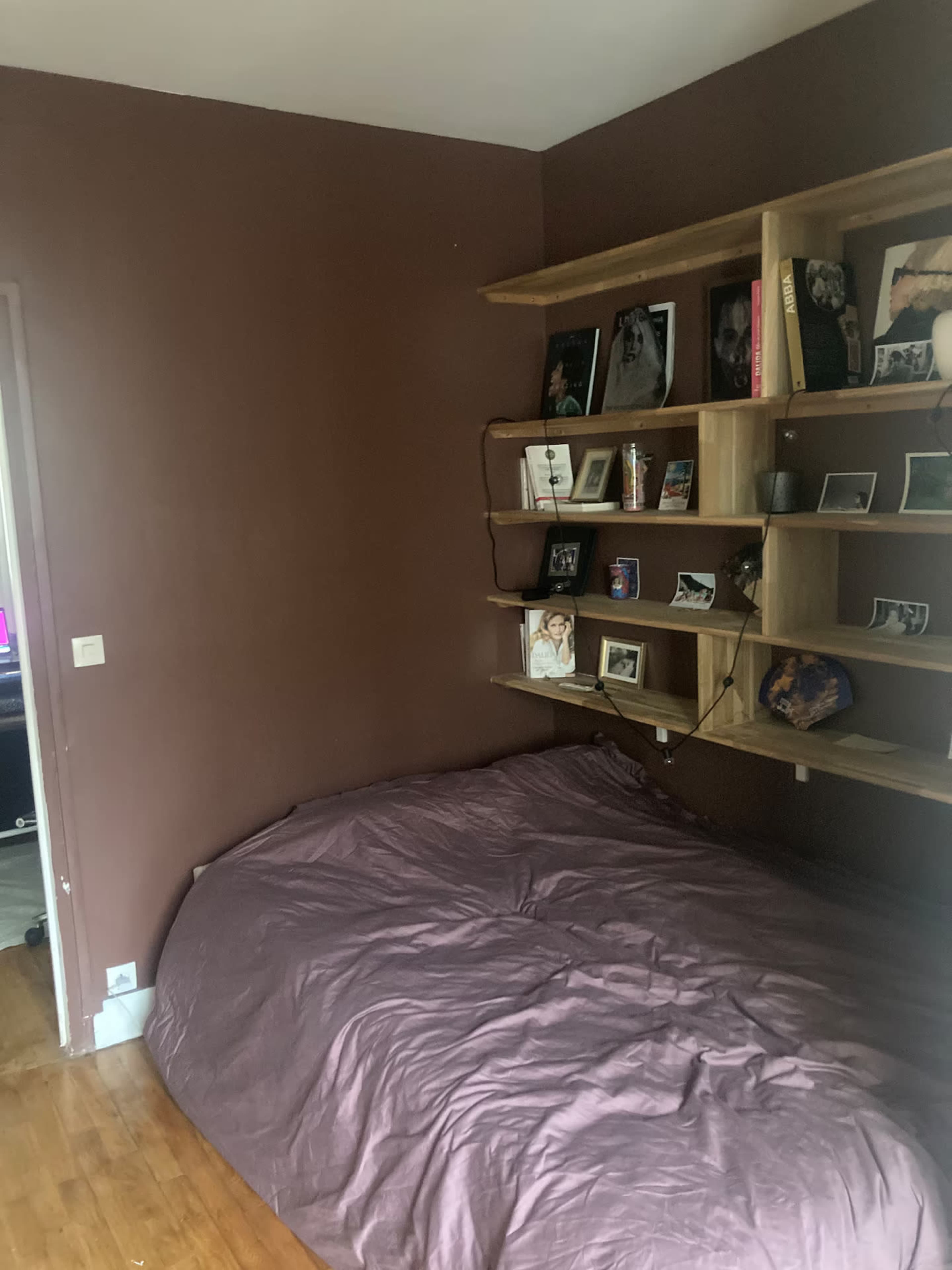 The image shows a small room featuring a brown wall, a large bed with a wrinkled purple cover, and a wooden shelf displaying various photographs and items.